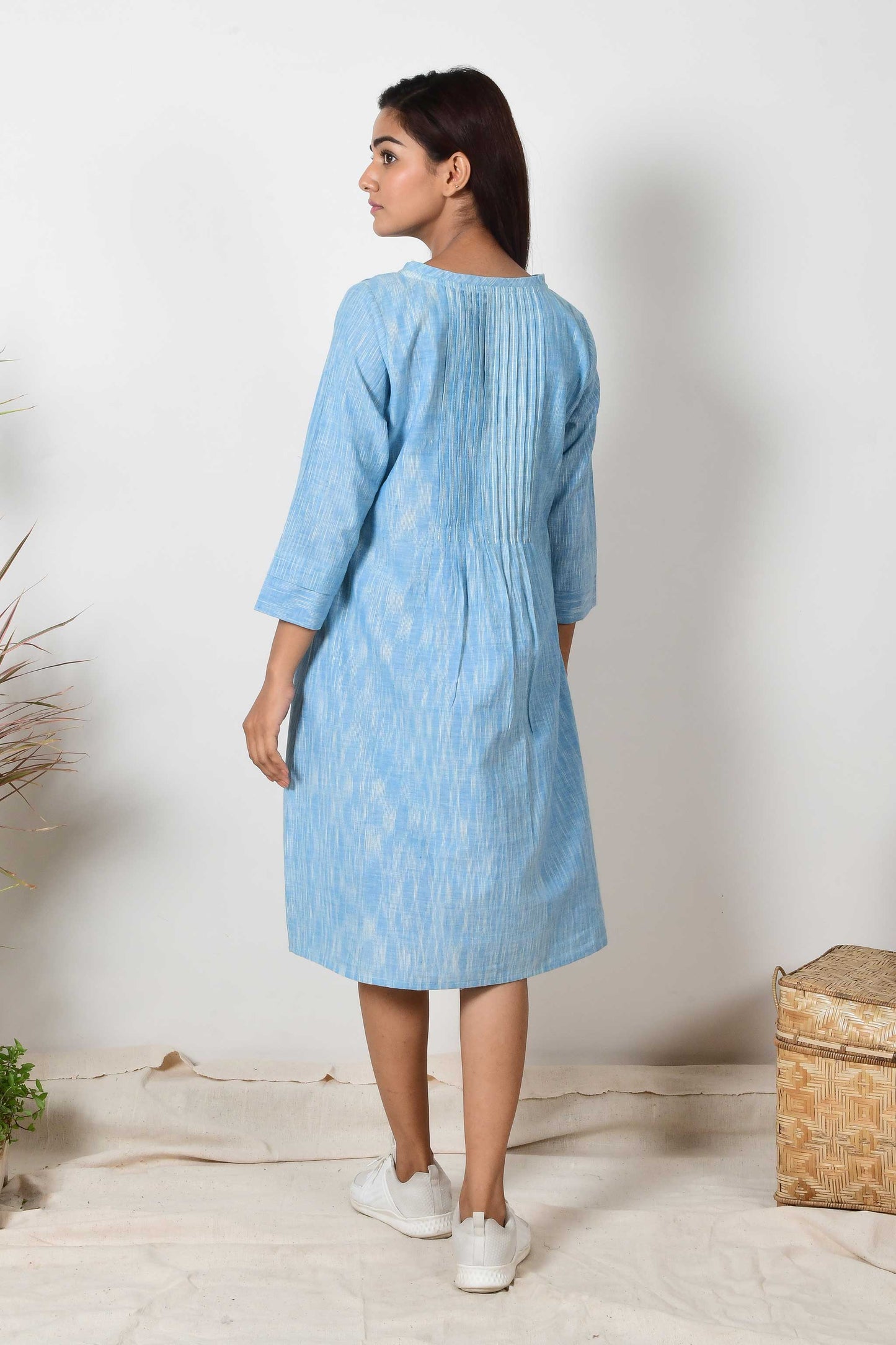 back of a sky blue cotton dress with pleated details worn by an Indian girl. Dress is paired with white sneakers.
