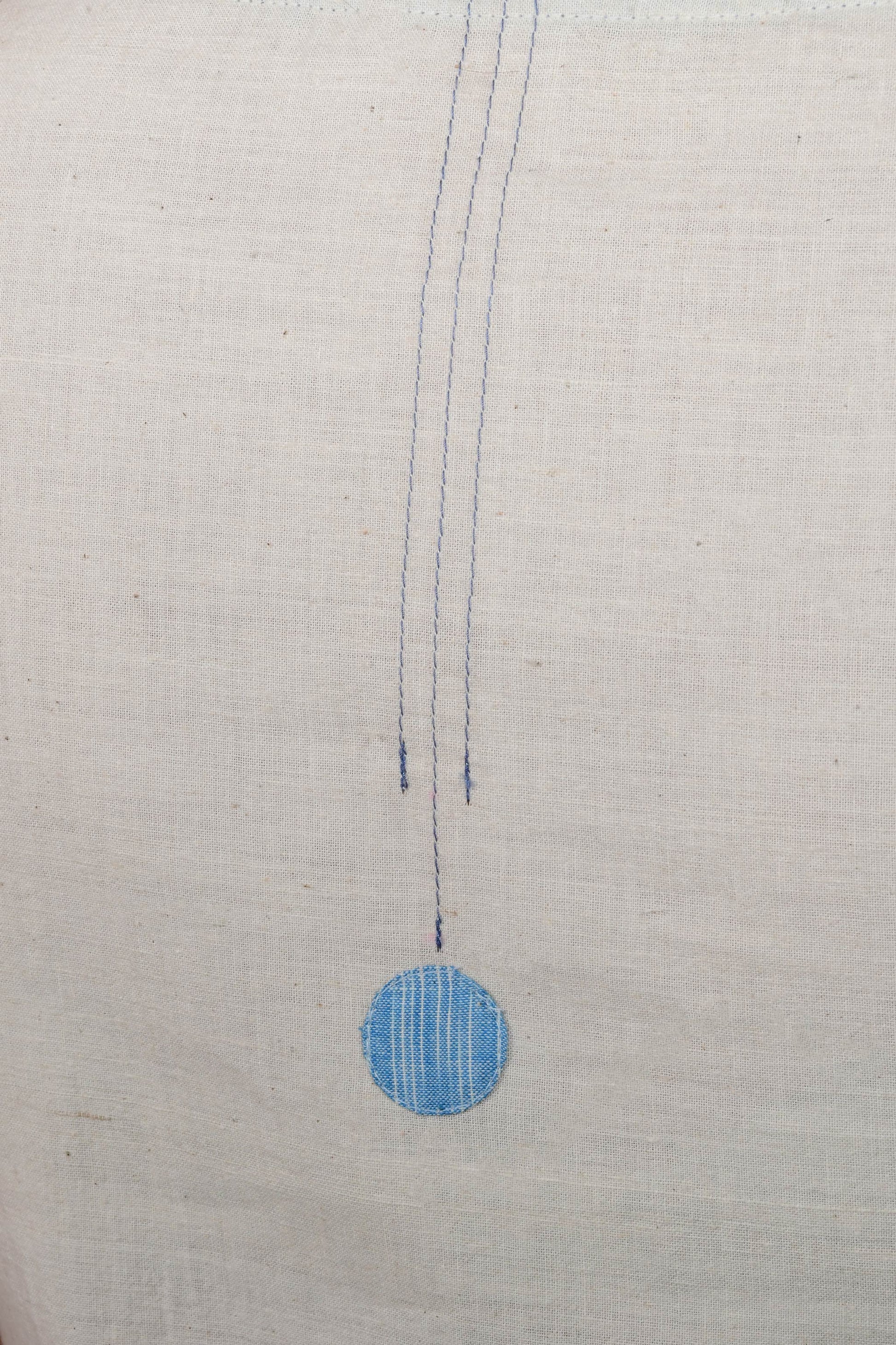 close up of a stitch detail and a blue circular patch on an off white cotton fabric base.