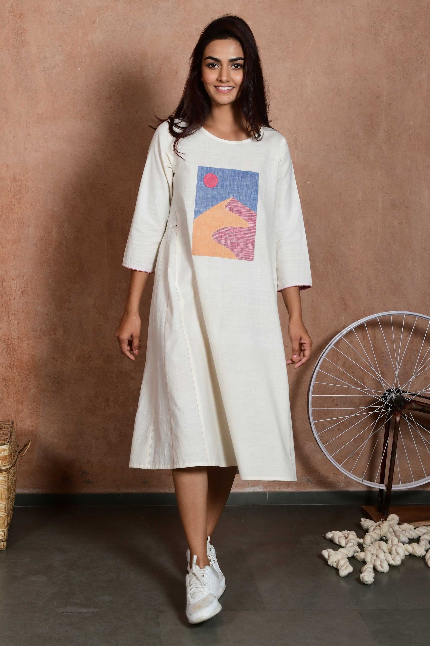 Front pose of a young indian model wearing a knee length off white dress with sleeves and an applique patch depicting a sand dune and sun.