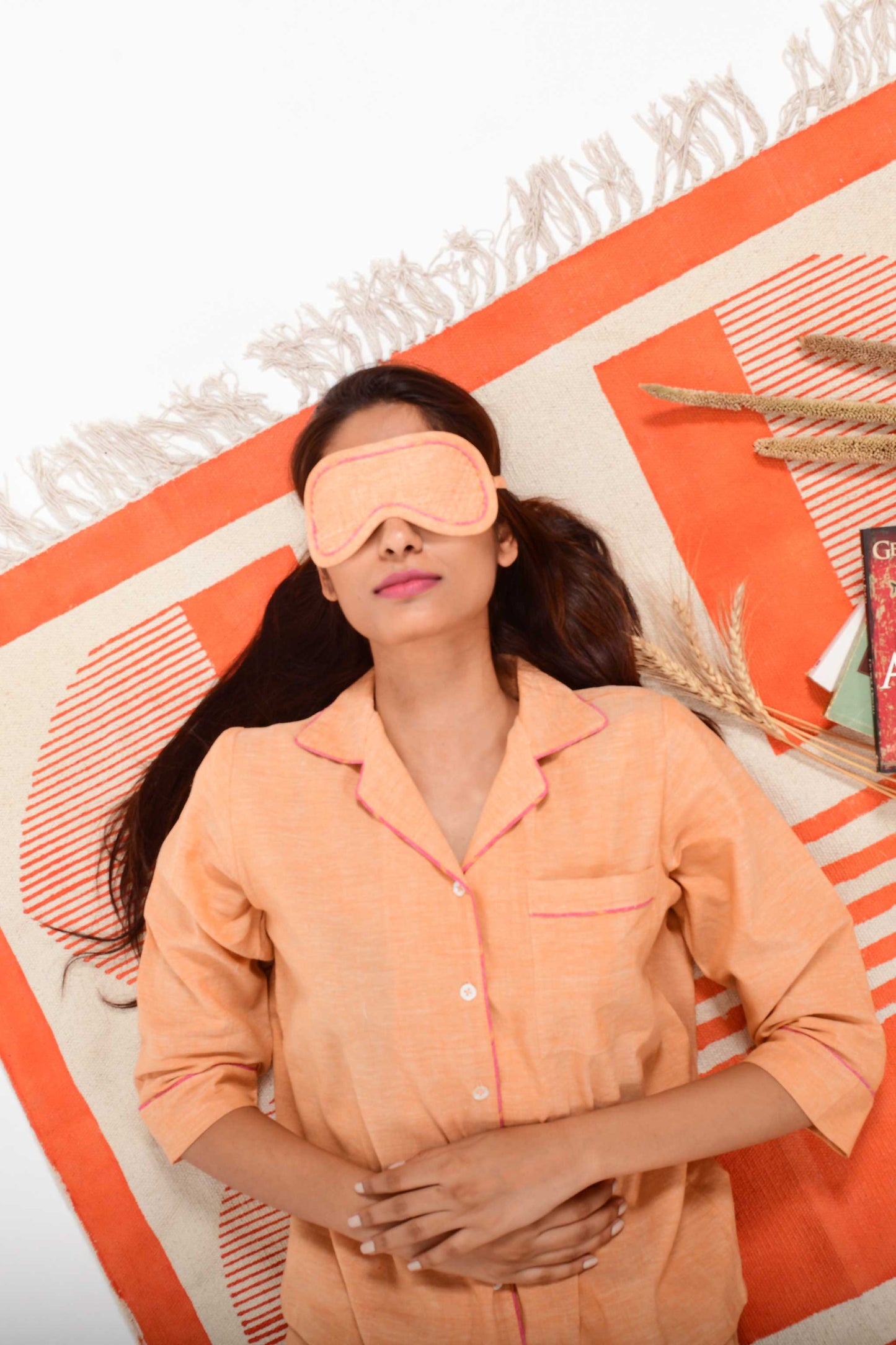 An Indian female womenswear fashion model wearing a orange chambray handspun and handwoven khadi cotton eye mask by Cotton Rack and lying on an orange carpet.