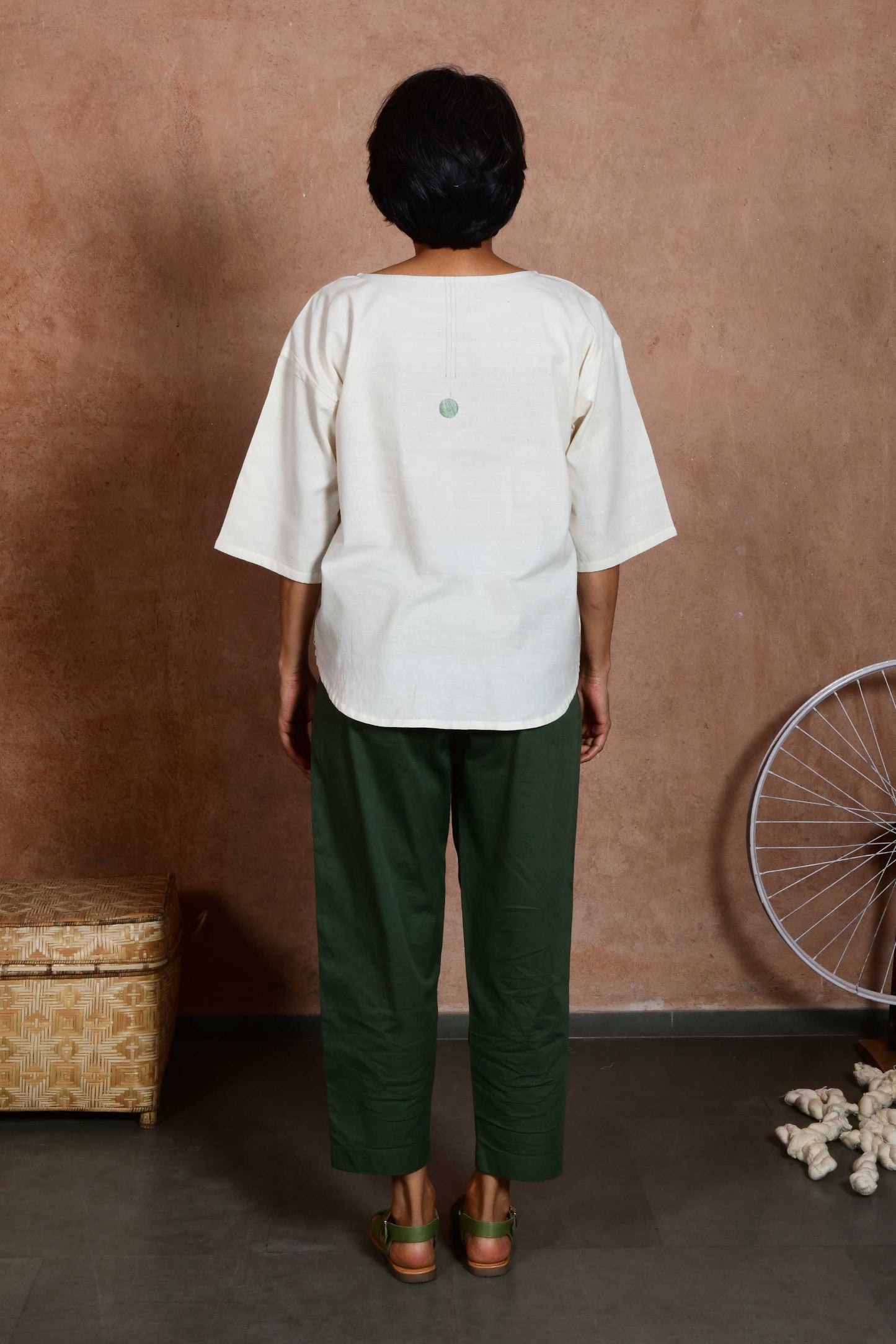Back pose a middle aged women wearing an off white oversized crop top blouse with an circular patchwork and stitch detail paired with green capri pants.