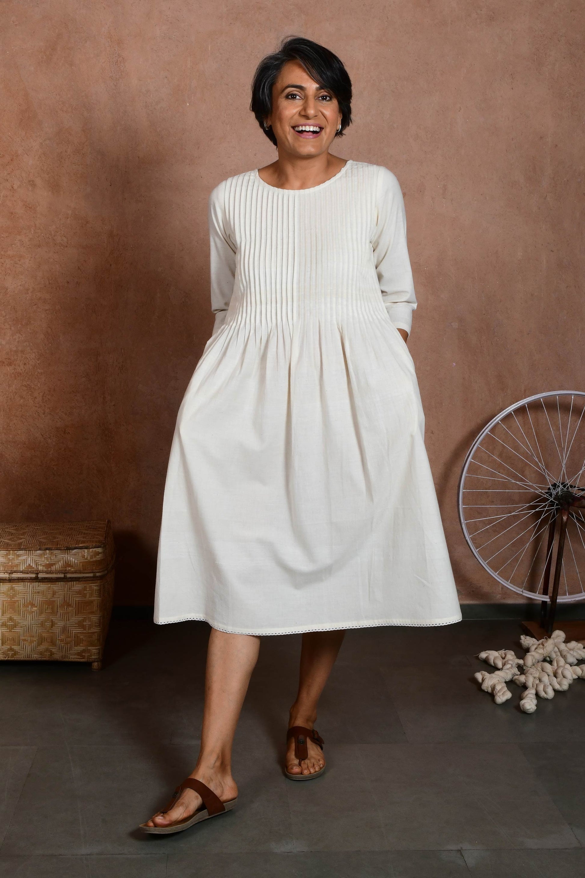 Front pose of a smilling middle aged indian woman with hands in pocket and wearing an off white knee length dress with sleeves and knife pleat detail.