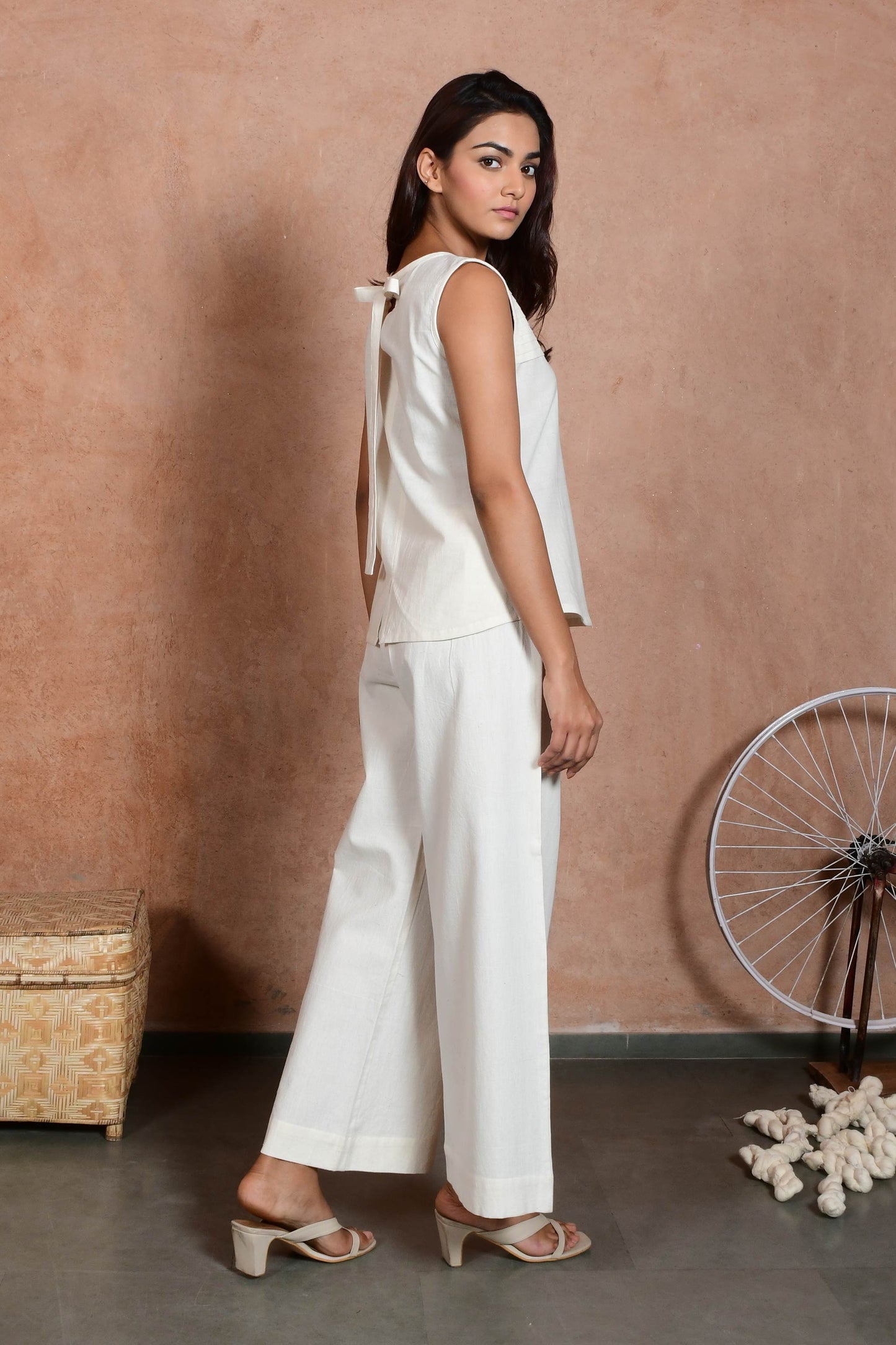 A young Indian model looking back posing wearing off white sleeveless pleated top at front with a bit of bow at the neck showing and straight pants.