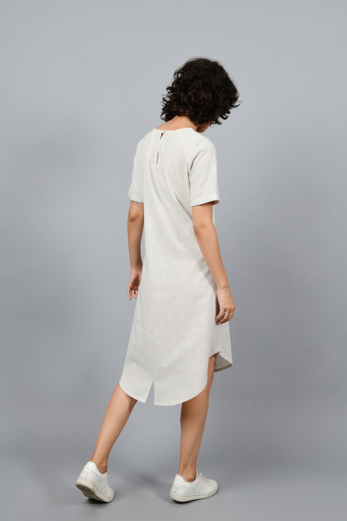 back of a womenswear fashion model wearing a minimally designed a-line round bottom western dress by Cotton Rack printed with blue stripes using brass block printing over handspun handwoven khadi cotton from maheshwar