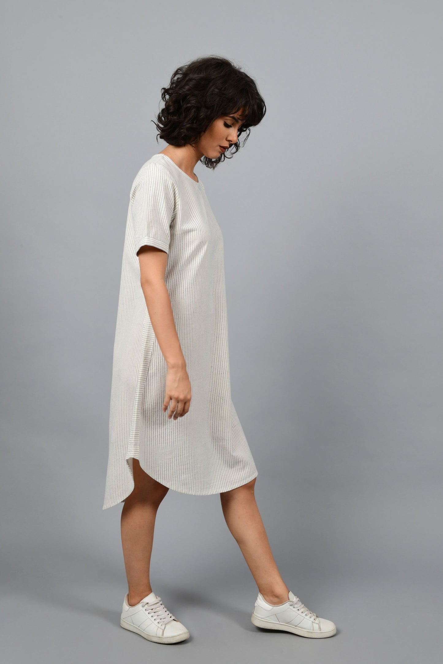 side pose of a womenswear fashion model wearing a minimally designed a-line round bottom western dress by Cotton Rack printed with blue stripes using brass block printing over handspun handwoven khadi cotton from maheshwar