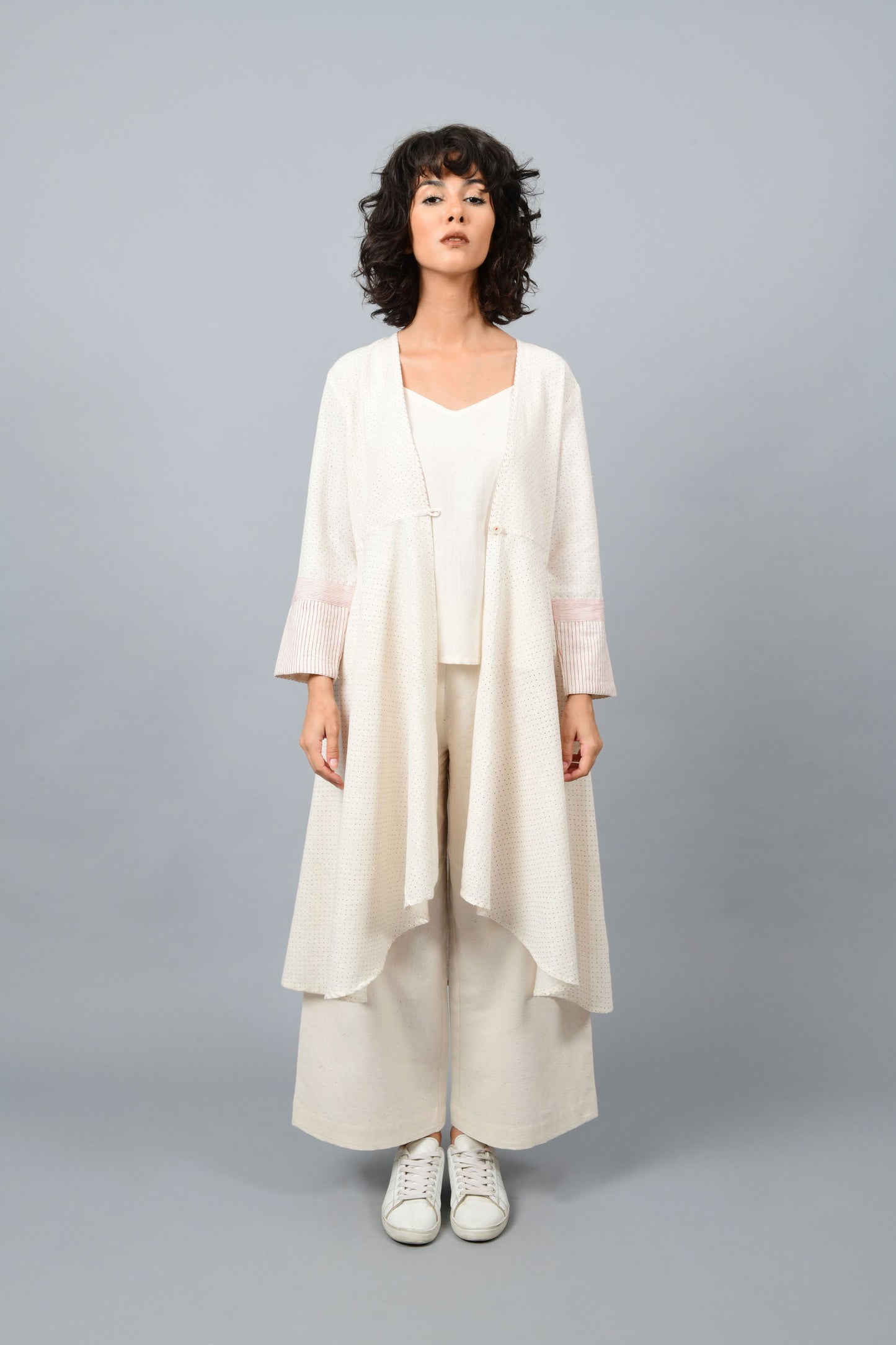 Model wearing a long flared off-white, natural khadi cotton shrug, hand block printed with red dots and red stripes on the sleeve hem.