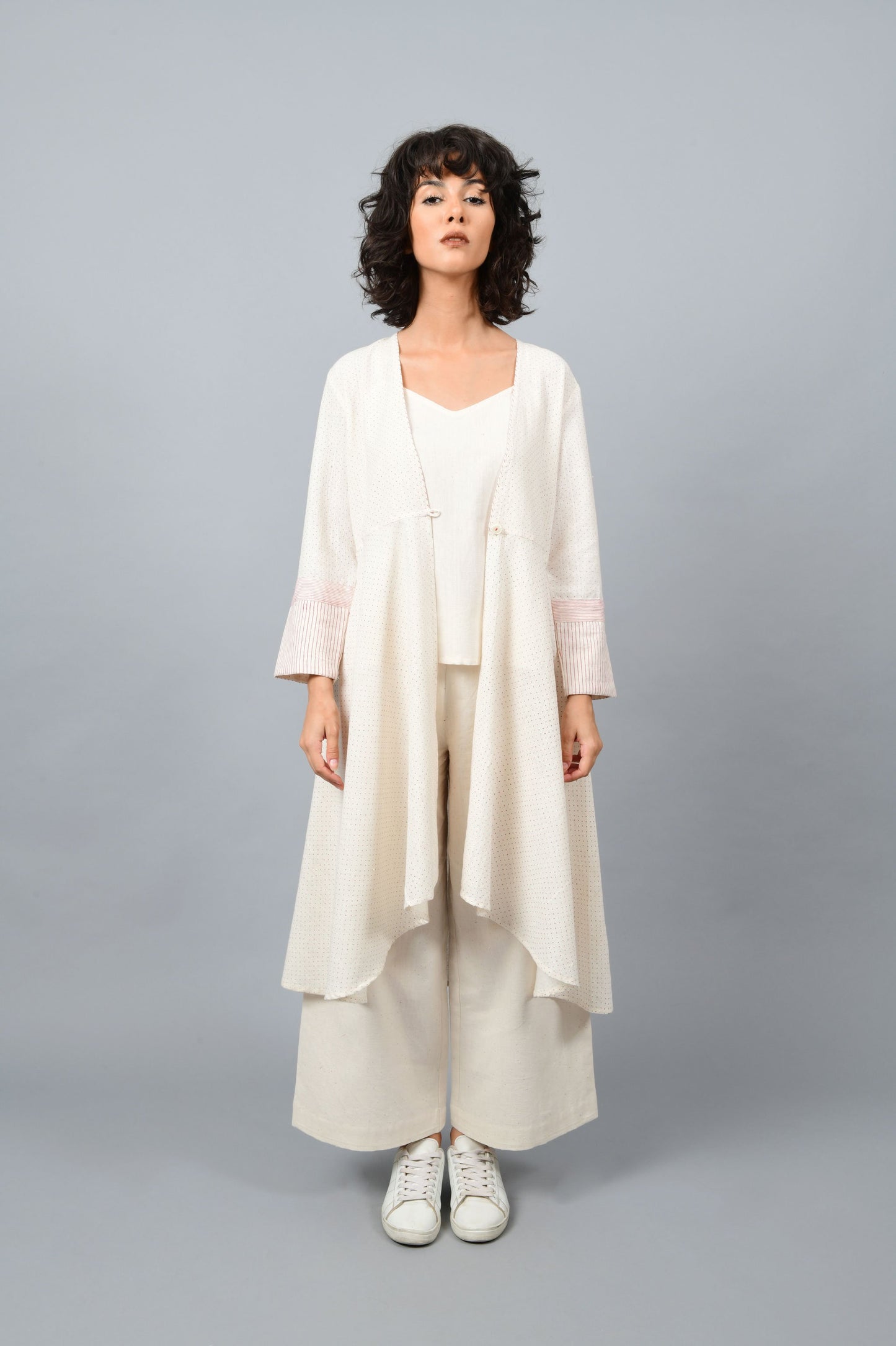 Model wearing a long flared off-white, natural khadi cotton shrug, hand block printed with red dots and red stripes on the sleeve hem.