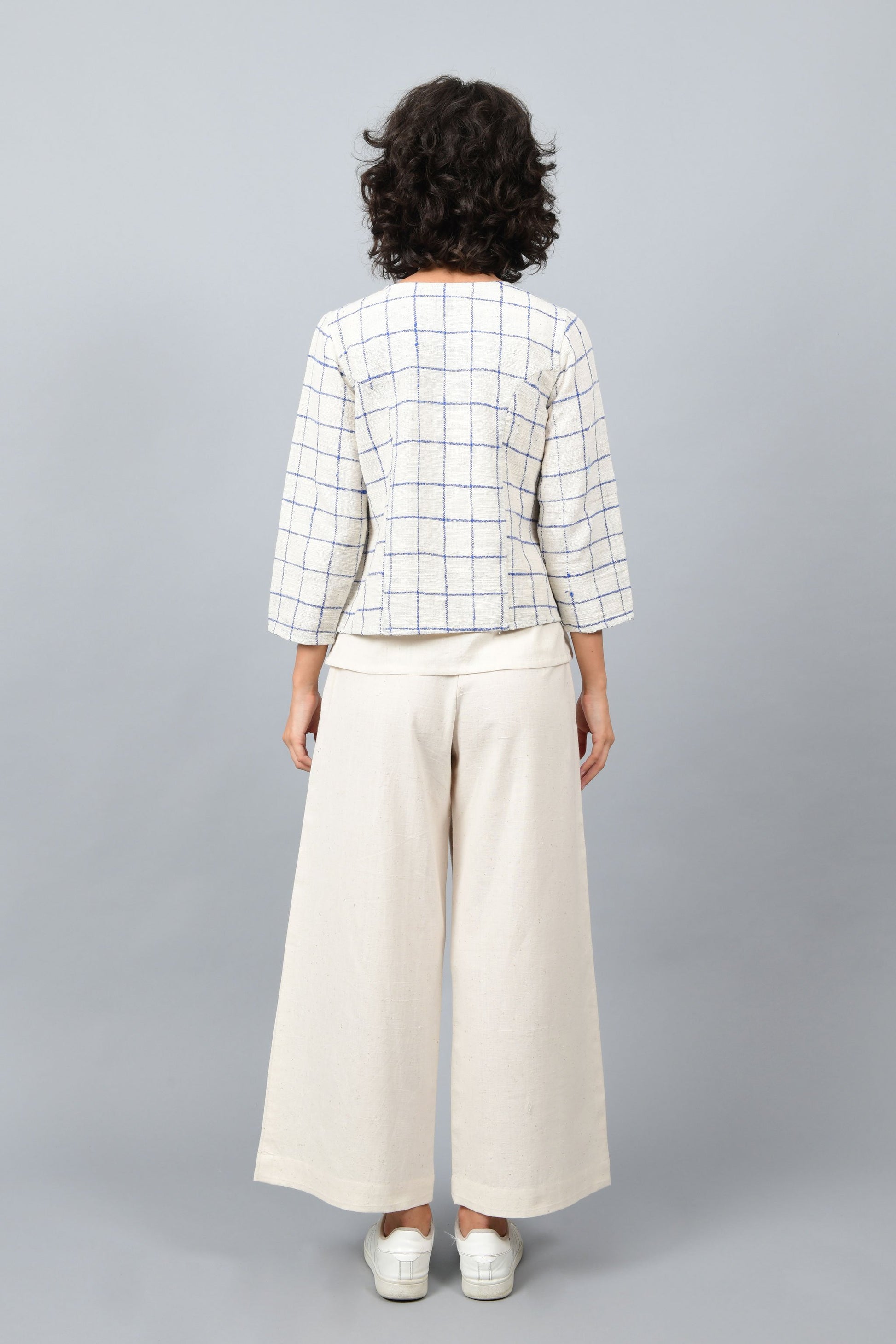 Model showing the back of open short jacket in thicker white handspun and handwoven khadi cotton with big blue checks over off-white spaghetti top and off-white palazzos paired with white sneakers.