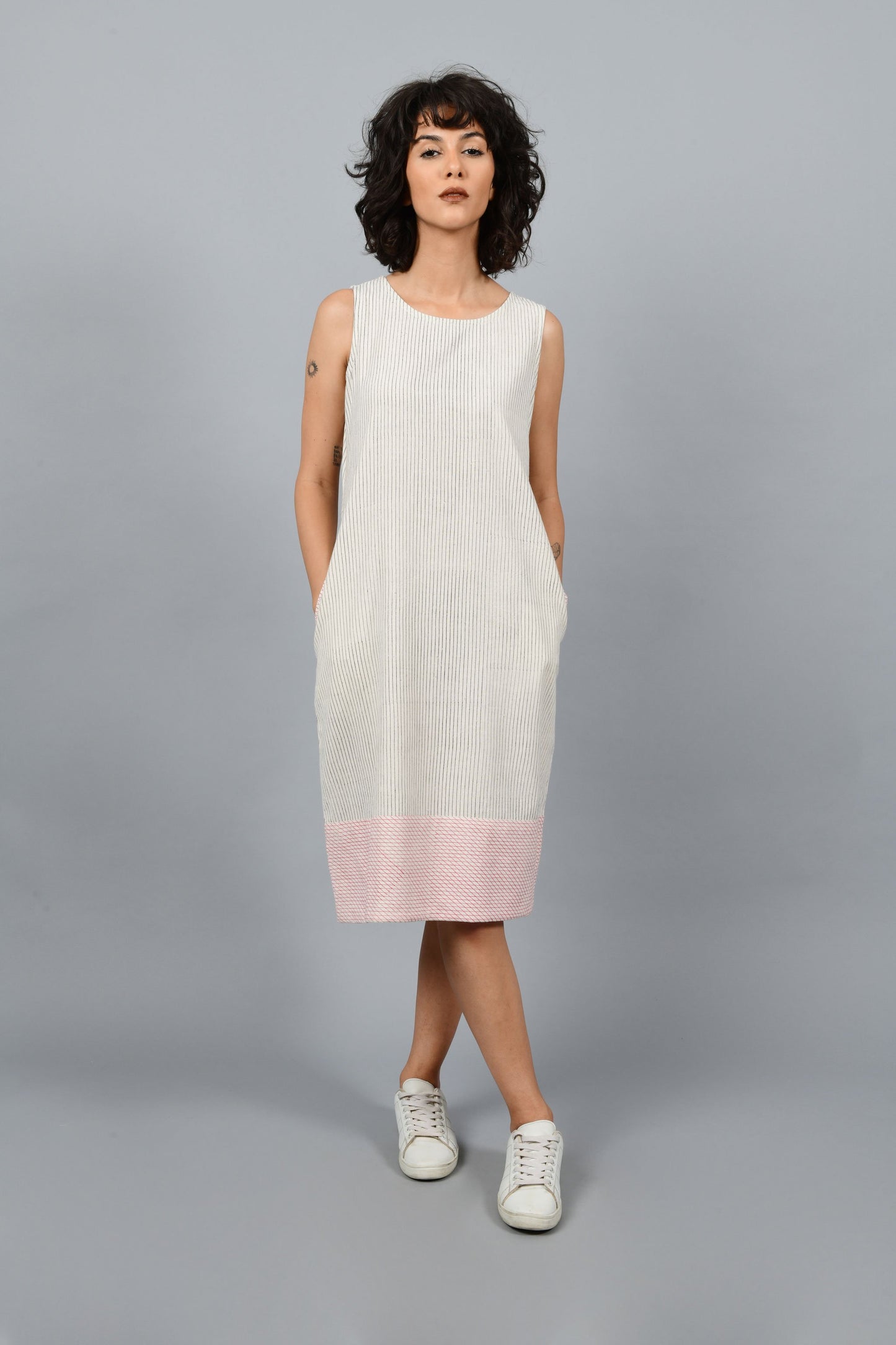 Model wearing black block printed black pin stripes with red checks on the hem on off-white handspun and handwoven khadi cotton sleeveless dress, posing straight for the camera.