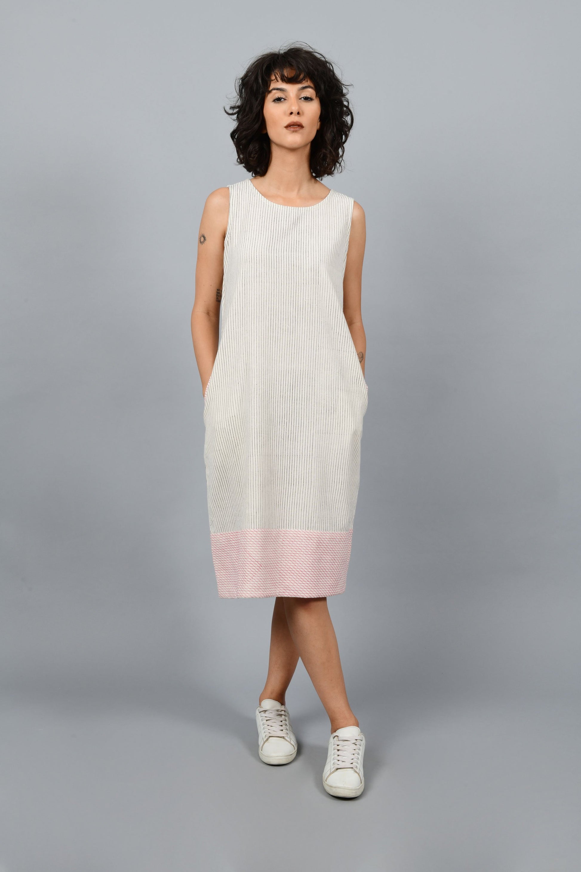 Model wearing black block printed black pin stripes with red checks on the hem on off-white handspun and handwoven khadi cotton sleeveless dress, posing straight for the camera.