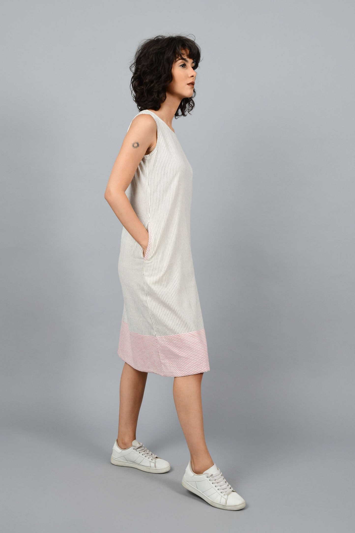Model wearing black block printed black pin stripes with red checks on the hem on off-white handspun and handwoven khadi cotton sleeveless dress, posing sideway for the camera.