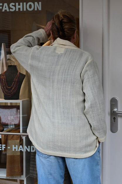 A middle aged european model looking inside the Via India Fashion Store in Netherlands Oosterbeek throug the glass wearing an ivory white woolen cardigan shrug with long sleeves over blue washed denims. Shrug is made of handspun and handwoven Indian wool by Cotton Rack.