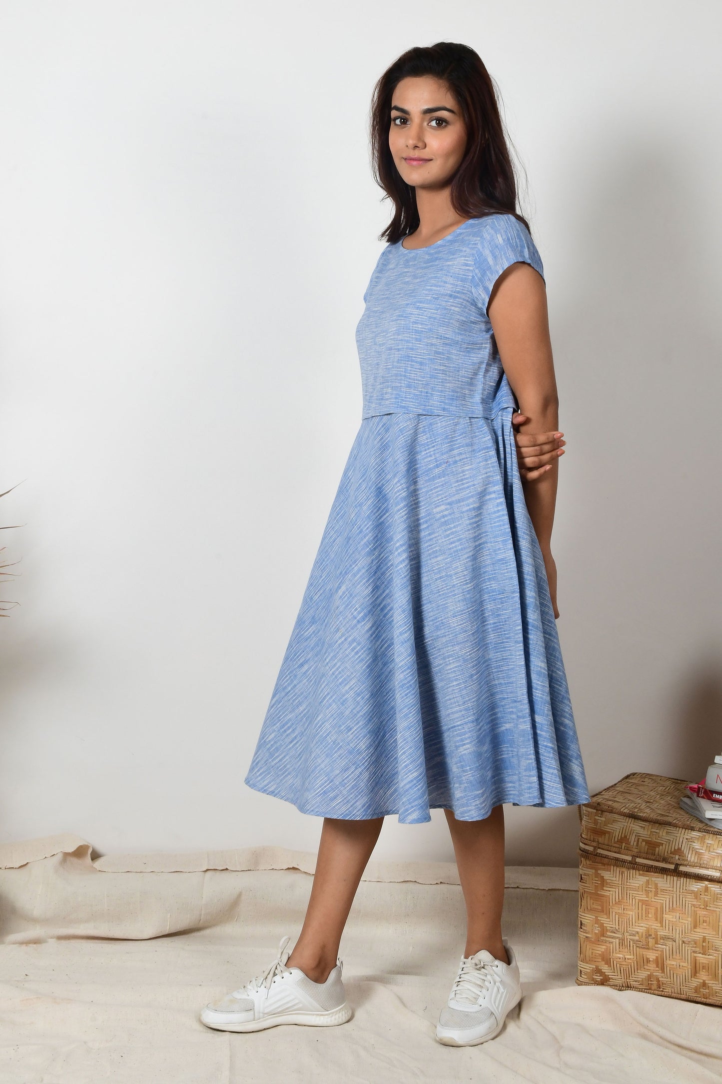 Indian girl with black hair and white sneakers wearing a blue a-line dress made with cotton that hand spun and hand woven giving a side pose as a model.