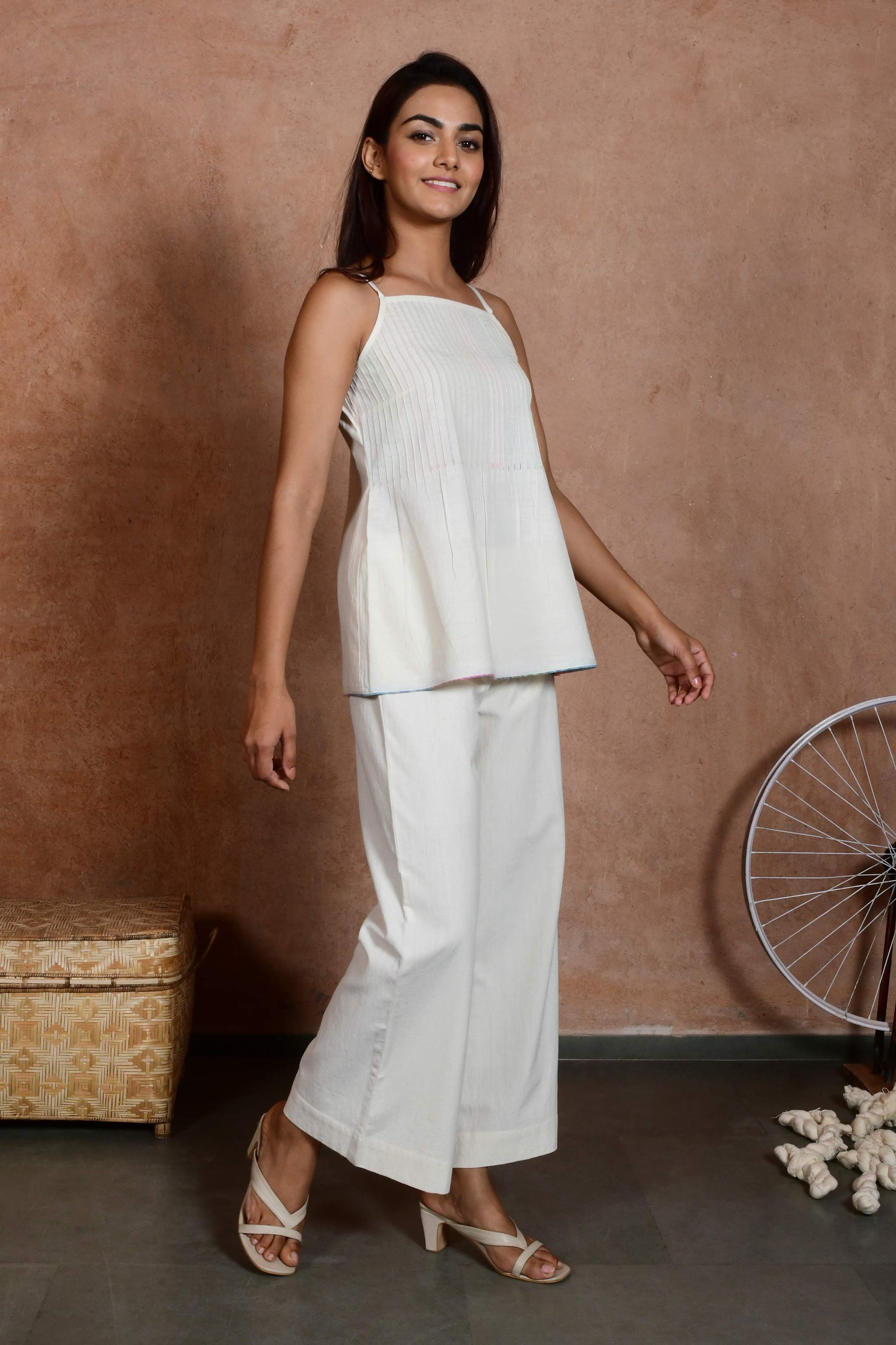 Three quarters pose of an indian model wearing an off white spaghetti top with pin tucks paired with off white straight pants.