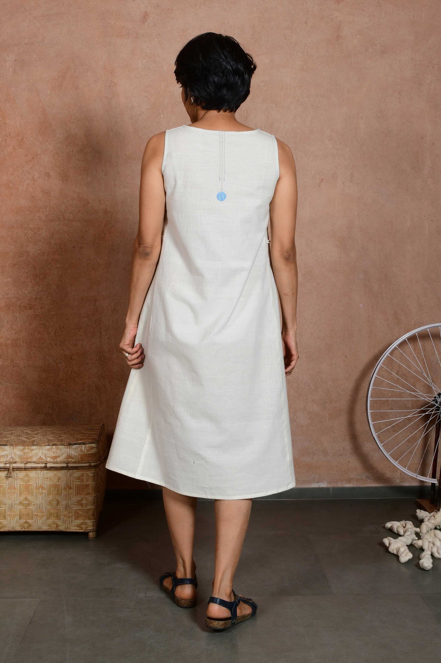 Back pose of a middle aged woman wearing a knee length off white dress made of handspun and handloom cotton fabric with a stitch detail and a small circular patch on the centre back of the neck.