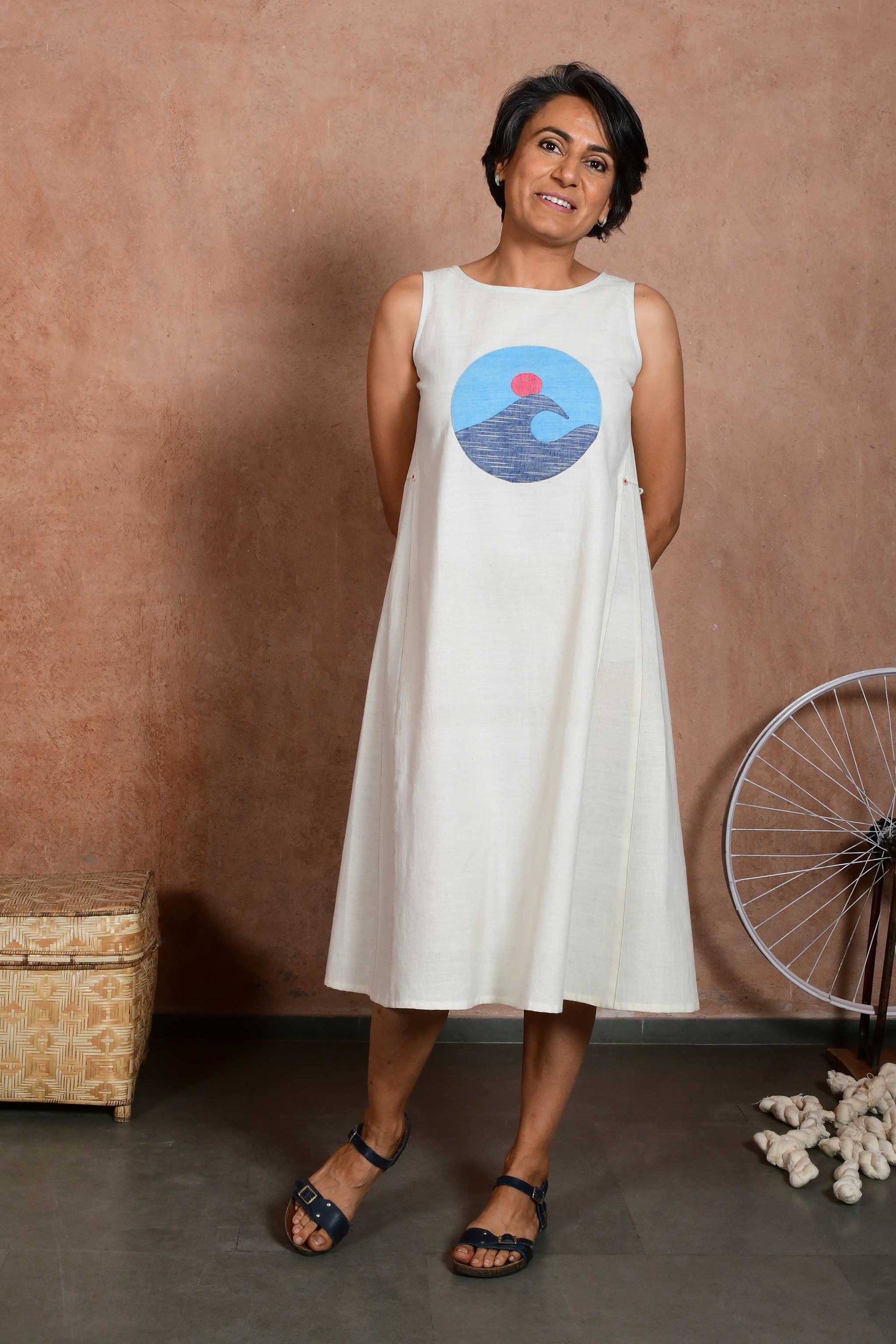 Front pose of a smiling middle aged woman wearing a knee length dress made of off white handspun handloom cotton fabric with a circular applique patch showing a wave in the sea with sun in the back.