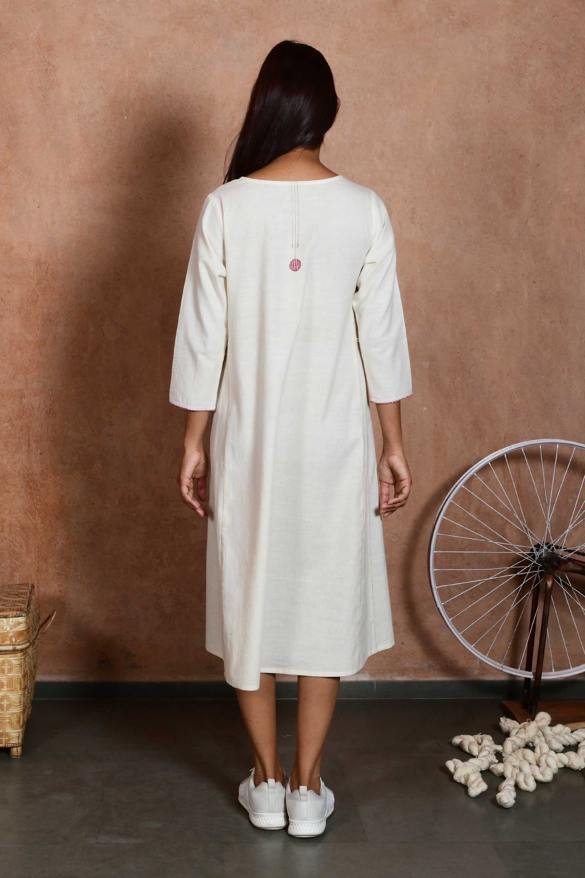 Back pose of a young indian model wearing a knee length off white dress with sleeves and a stitching detail on the back.