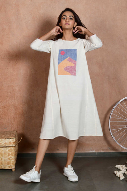 Front pose of a young indian model wearing a knee length off white dress with sleeves and an applique patch depicting a sand dune and sun.