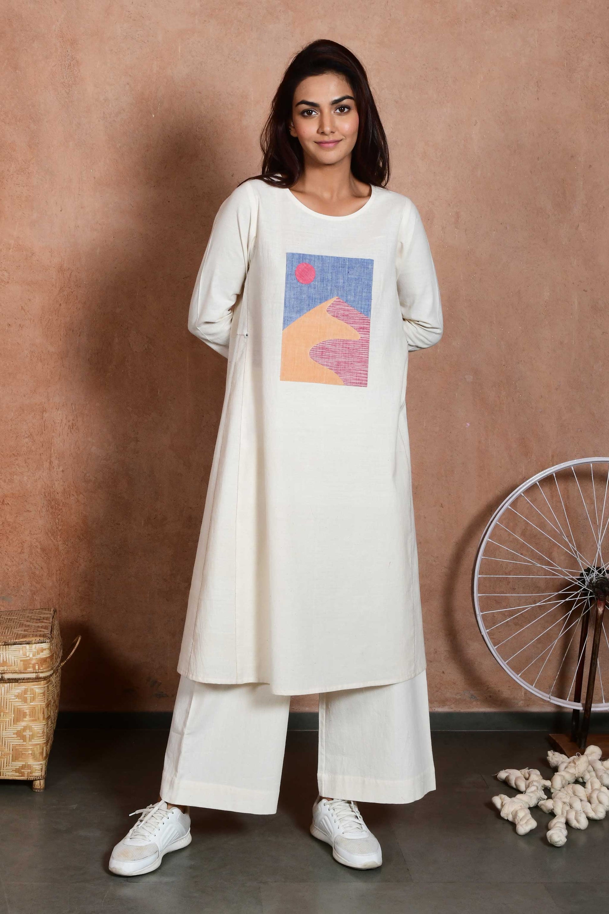 Front pose of a young indian model wearing a knee length off white dress with sleeves and an applique patch depicting a sand dune and sun.
