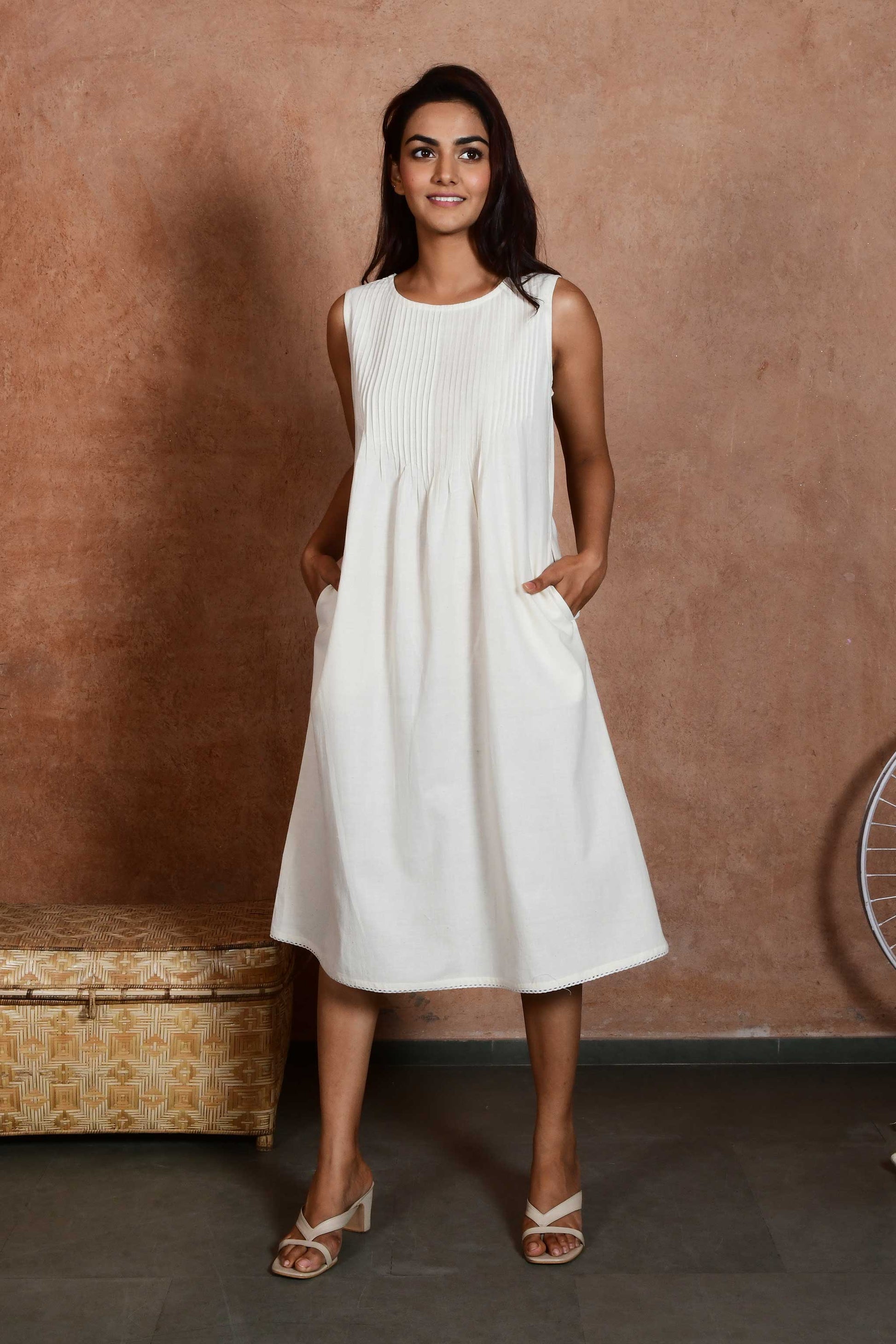 Front pose of a young indian model wearing an off white knee length a line dress with pleats till waist with hands in pockets.