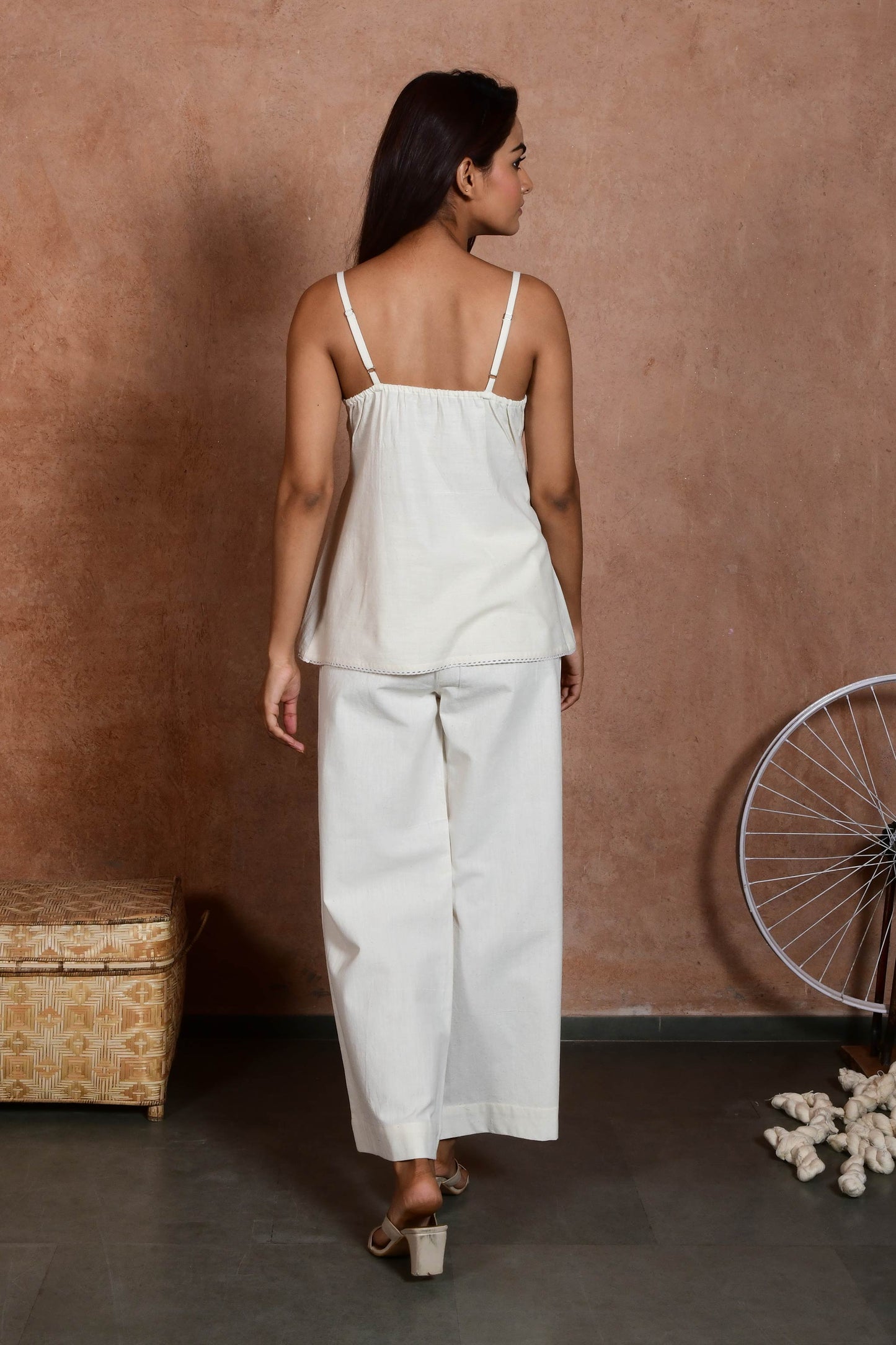 Back pose of an indian model wearing an off white spaghetti top with pin tucks paired with off white straight pants.