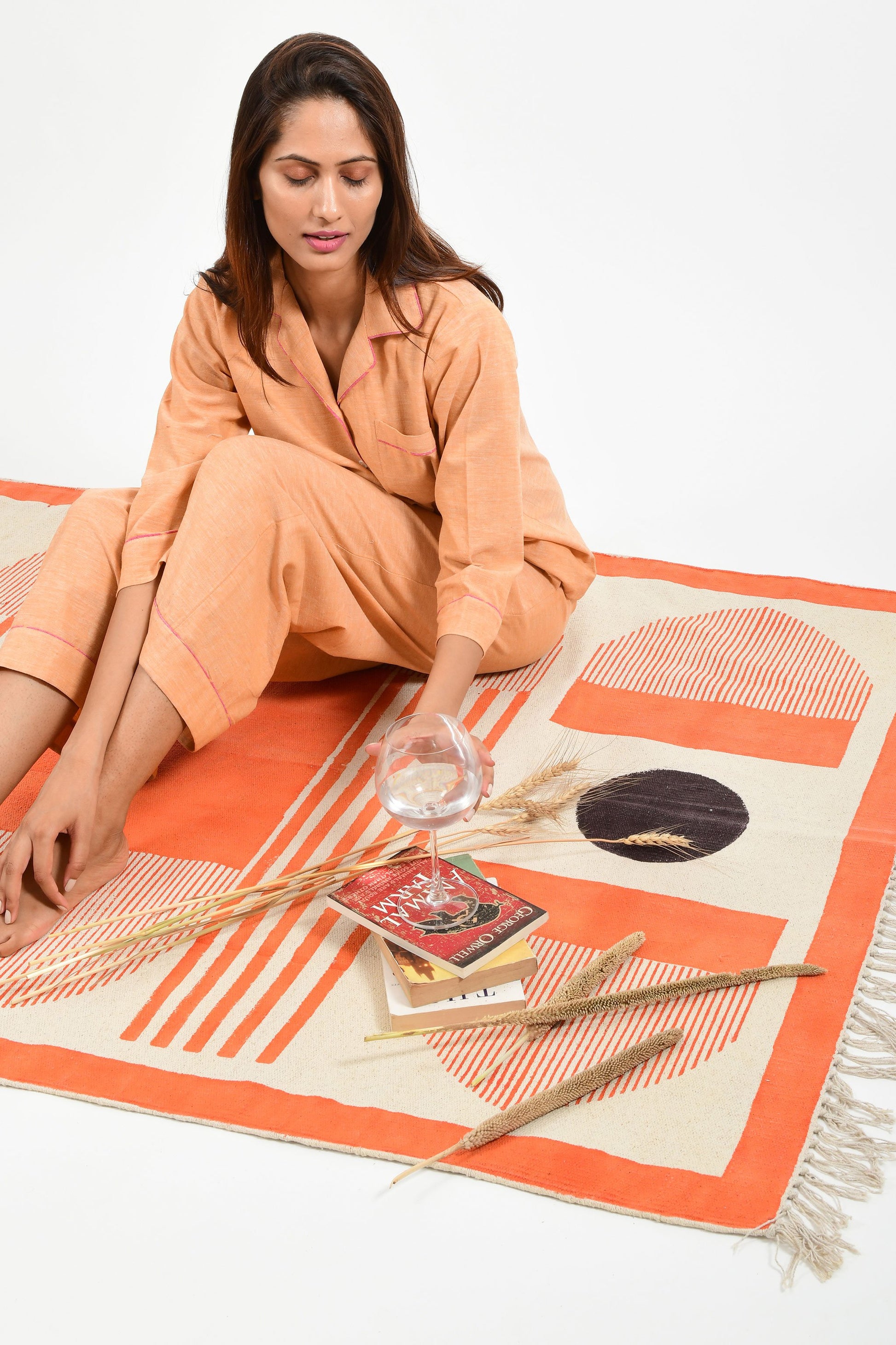 Creative pose of an Indian female womenswear fashion model in azo-free dyed handspun and handwoven khadi cotton nightwear pyjama & shirt in orange chambray by Cotton Rack and sitting on a handmade carpet with books and wine glass filled with water.