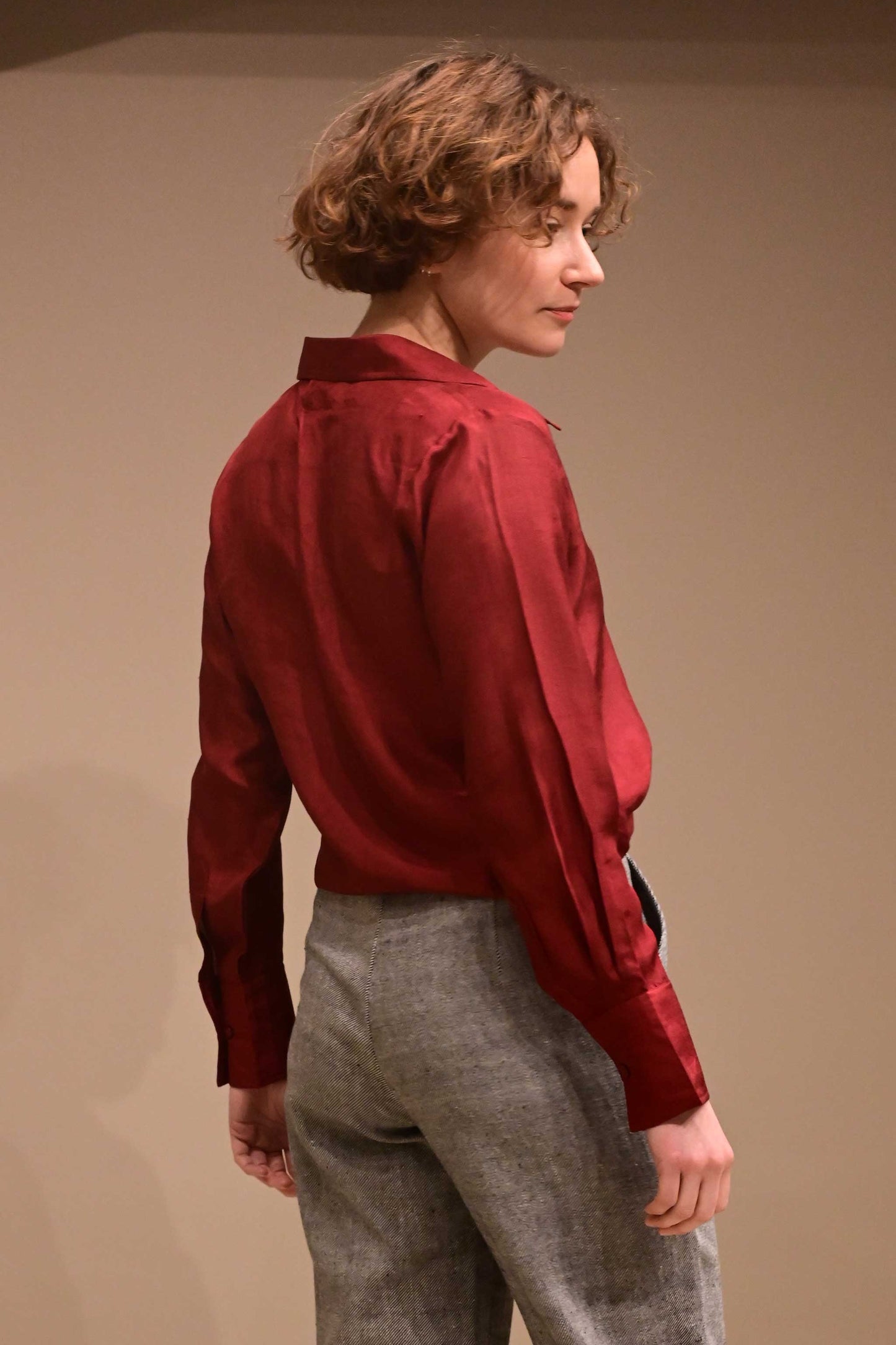 Back profile of a caucasian female model wearing a collared silk shirt in wine red color that is made from ethically sourced hand reeled and hand spun Indian silk.