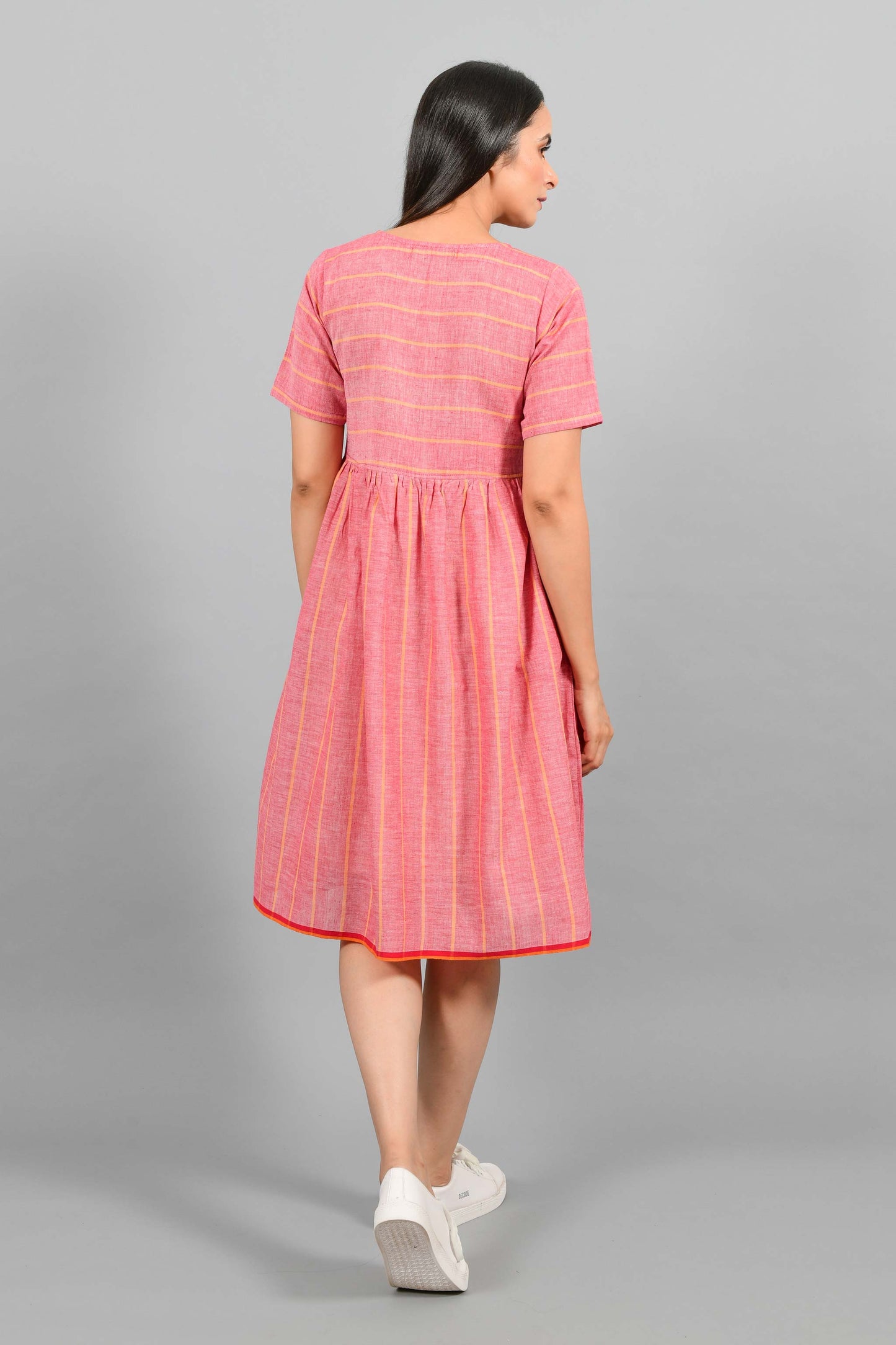 Back pose of an Indian female womenswear fashion model in a red chambray handspun and handwoven khadi cotton with orange stripes by Cotton Rack. The dress has front buttoned yoke and gathers at waist.