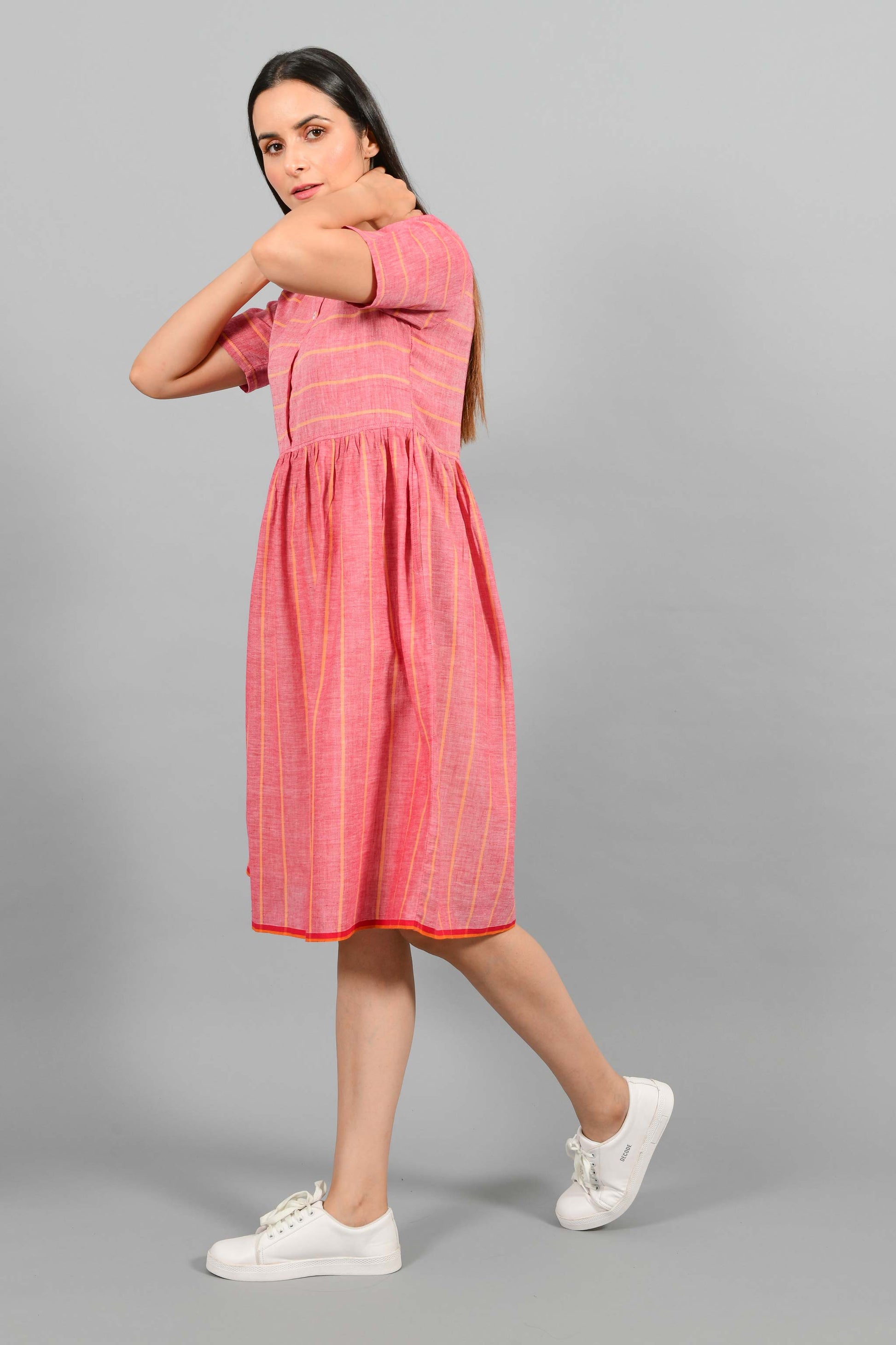 Side walking pose of an Indian female womenswear fashion model in a red chambray handspun and handwoven khadi cotton with orange stripes by Cotton Rack. The dress has front buttoned yoke and gathers at waist.