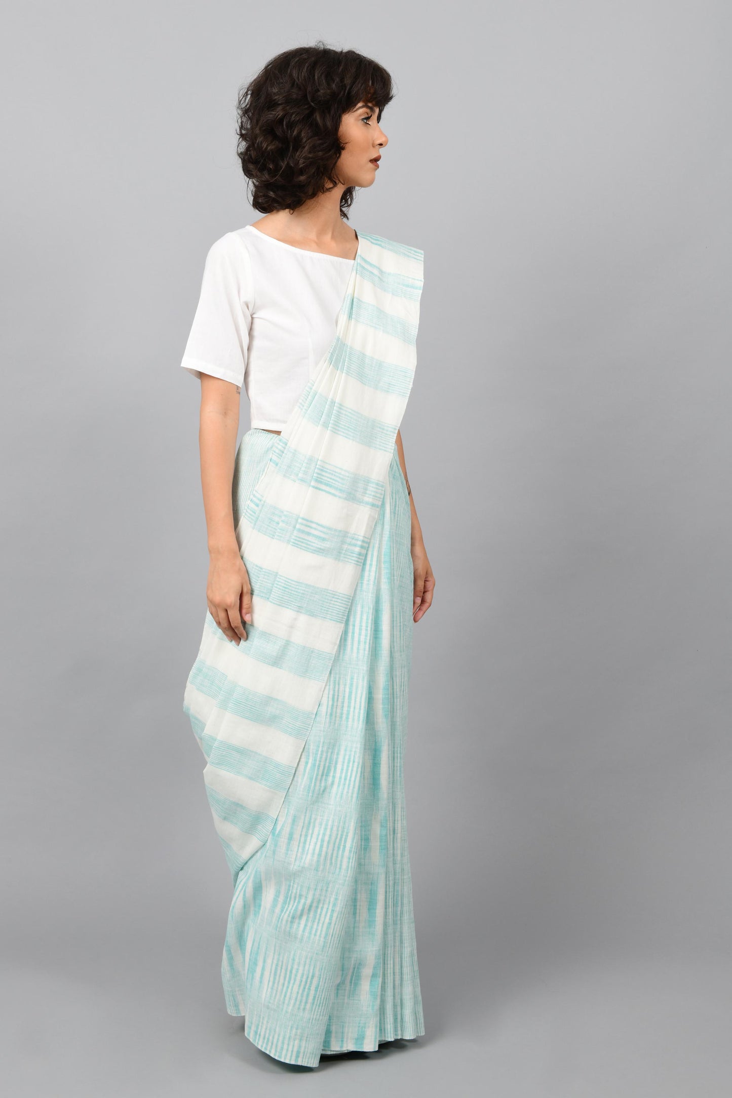 Side pose of a female womenswear fashion model draped in a aquamarine blue & white space dyed homespun and handwoven cotton sari by Cotton Rack.