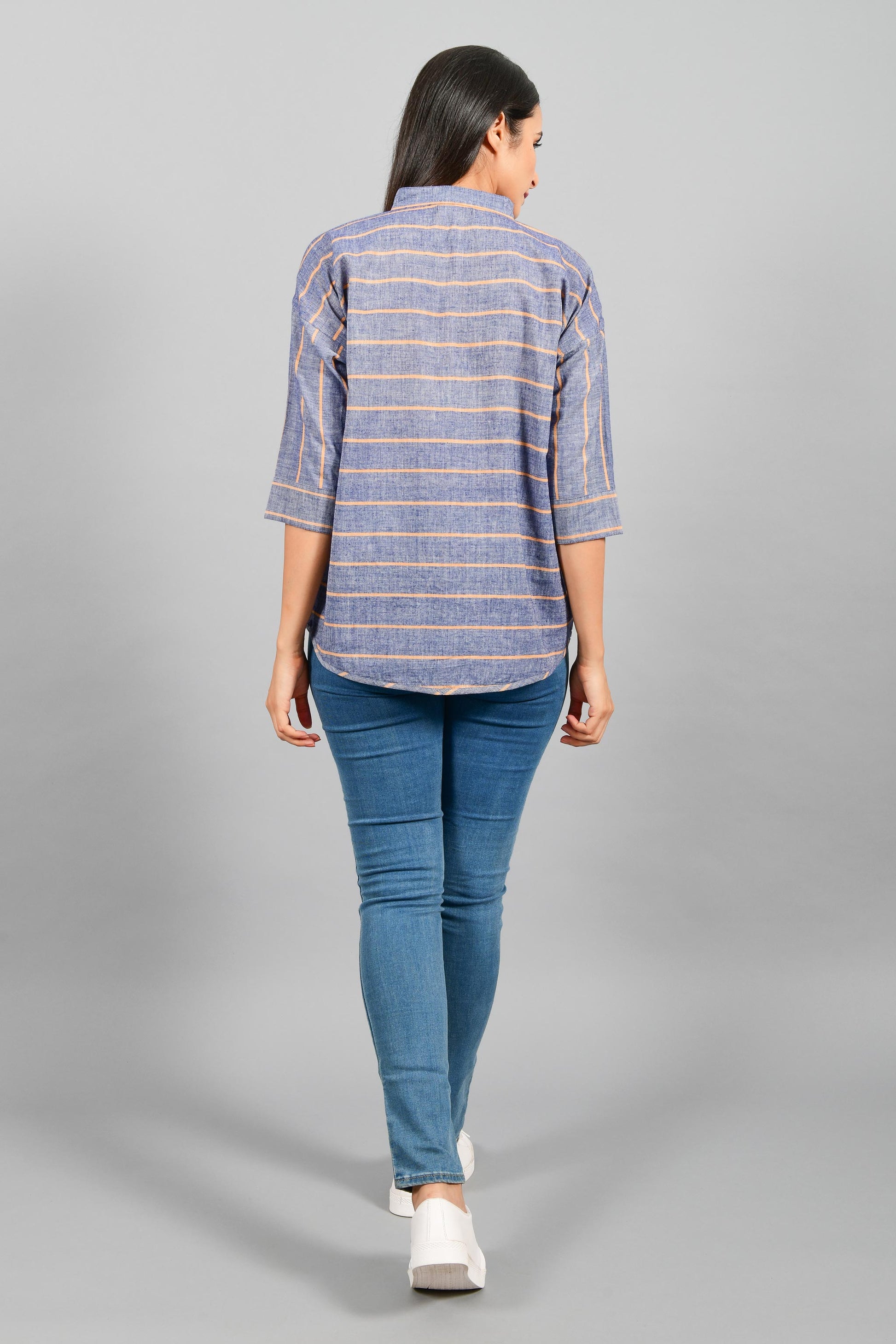 Back pose of an Indian female womenswear fashion model in a blue chambray with orange stripes handspun and handwoven khadi cotton free size top by Cotton Rack.