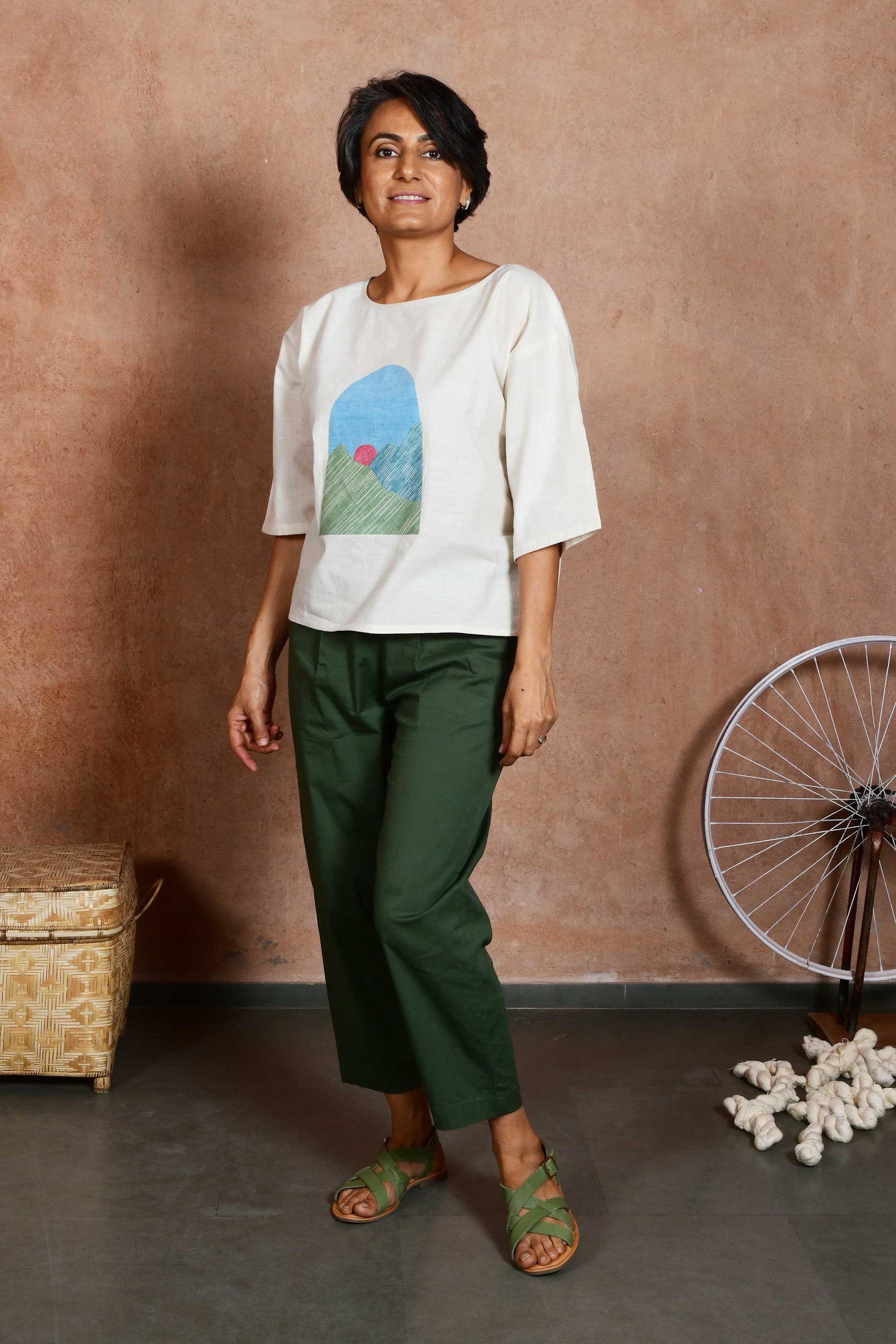 Front three quarters pose of a middle aged women wearing an off white oversized crop top blouse with an applique patch depicting a sunrise and green mountains paired with green capri pants.