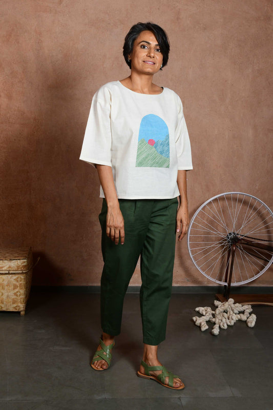Front pose of a middle aged women wearing an off white oversized crop top blouse with an applique patch depicting a sunrise and green mountains paired with green capri pants.
