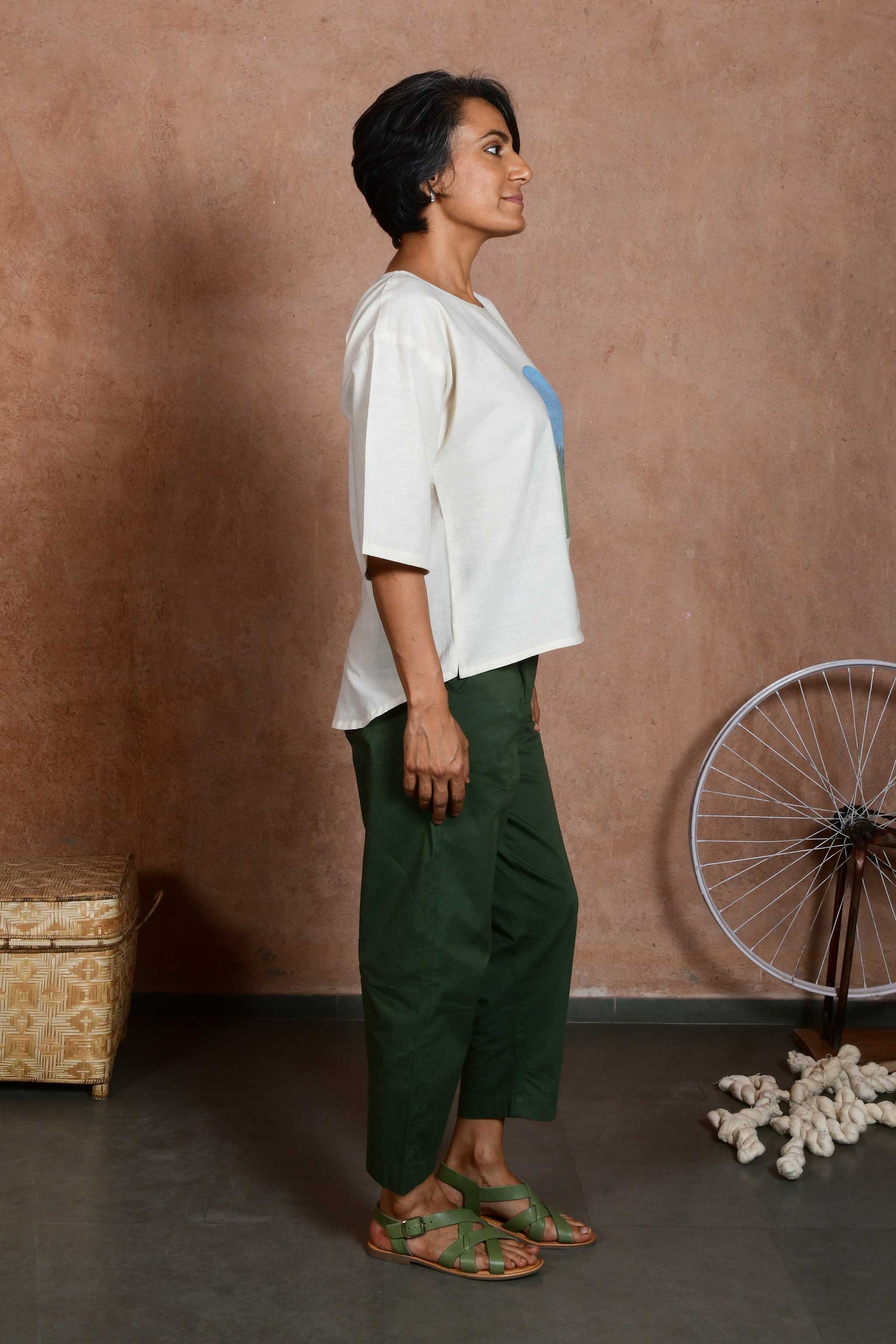 Side pose a middle aged women wearing an off white oversized crop top blouse with an applique patch depicting a sunrise and green mountains paired with green capri pants.