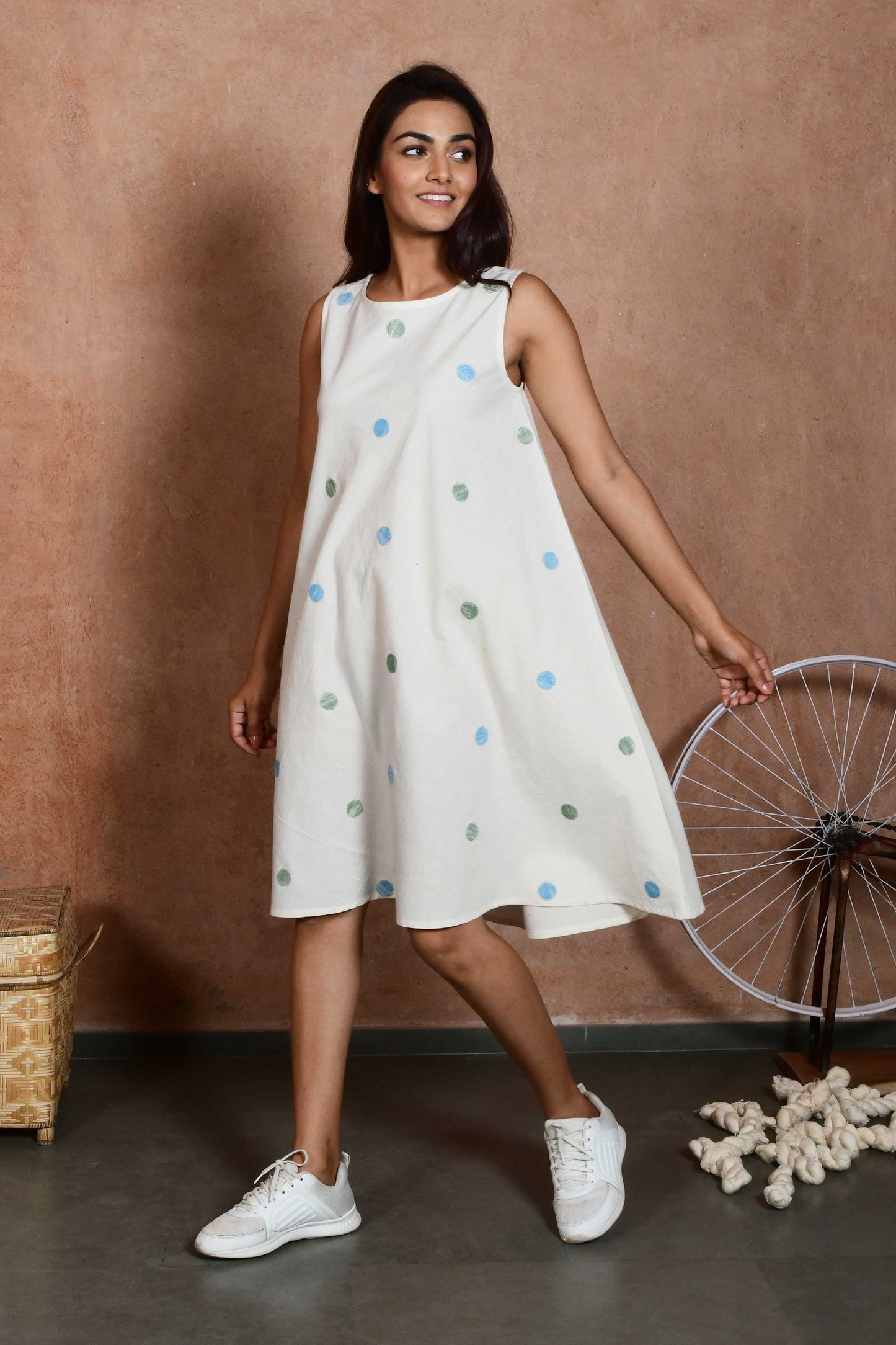 Side pose of an indian model wearing a handspun handloom cotton sleeveless dress that is knee length with green and blue polka dot patches.