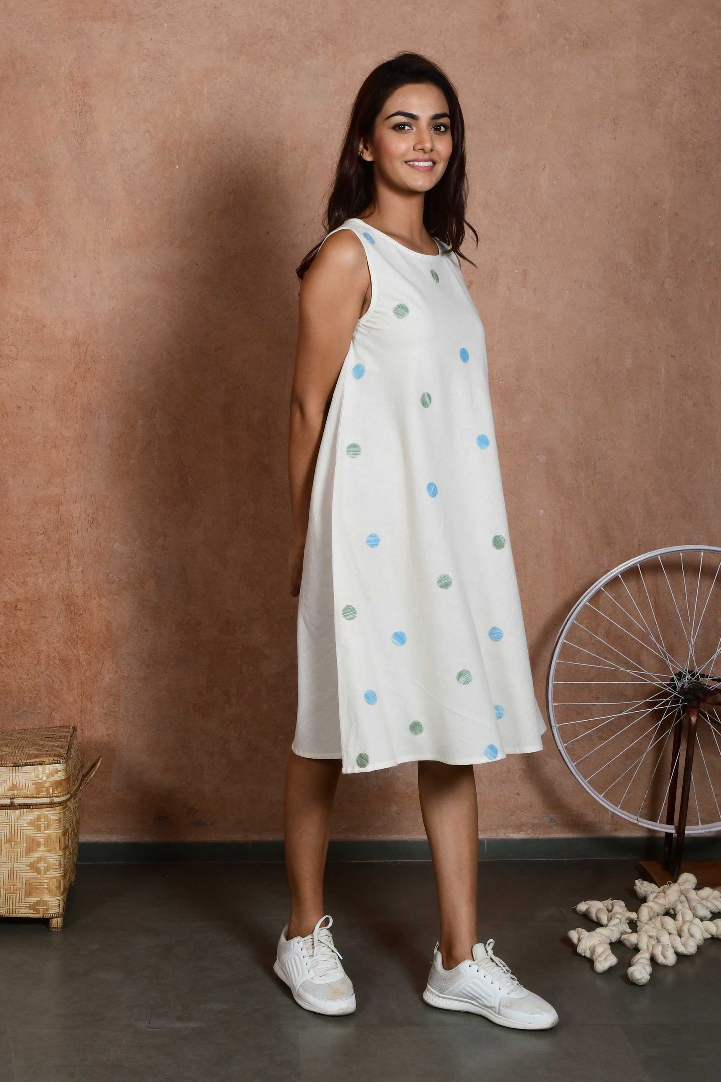 Three quarter pose of an indian model wearing a handspun handloom cotton sleeveless dress that is knee length with green and blue polka dot patches.