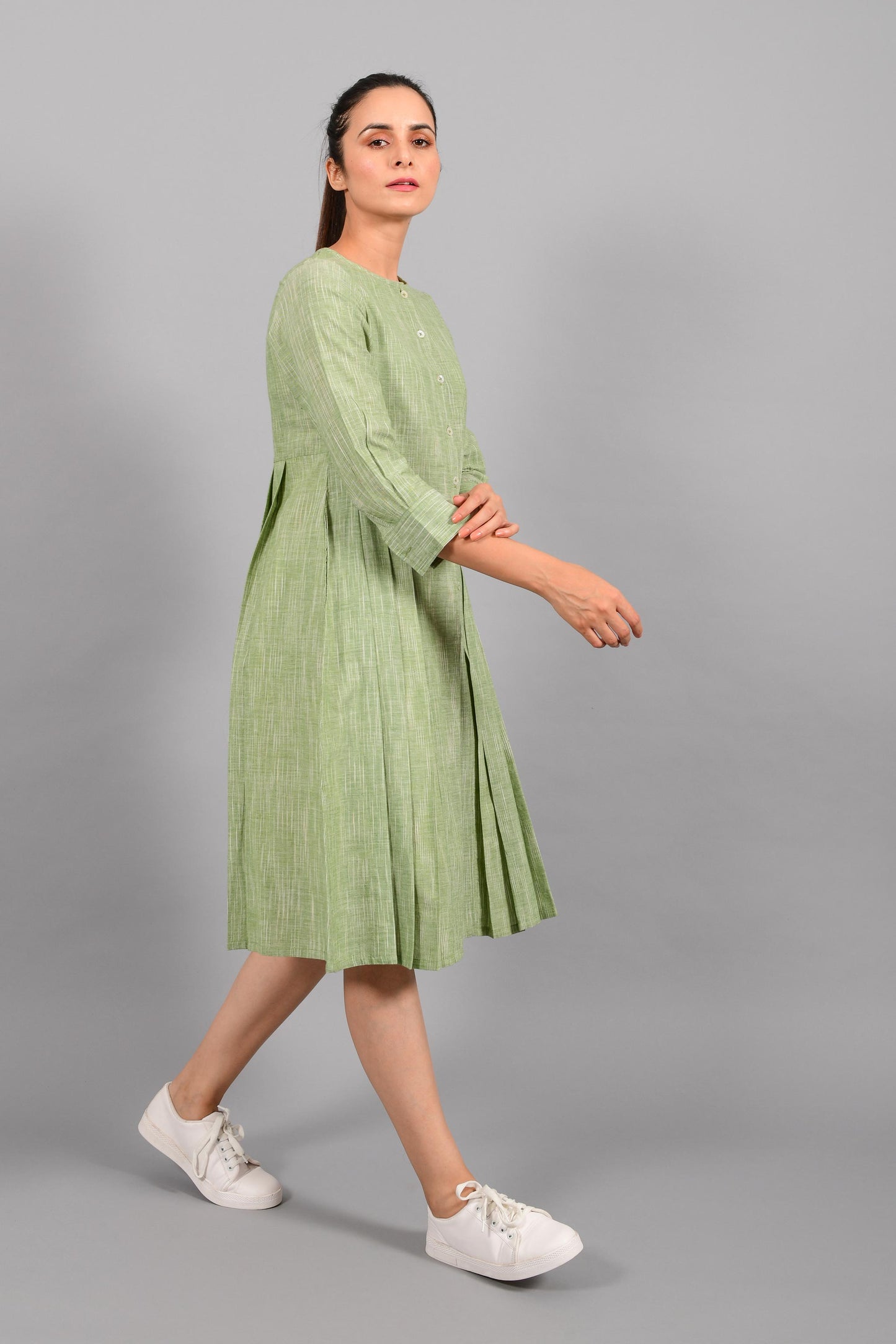 Stylised side pose of an Indian female womenswear fashion model in a space dyed olive green handspun and handwoven khadi cotton pleated dress-kurta by Cotton Rack.