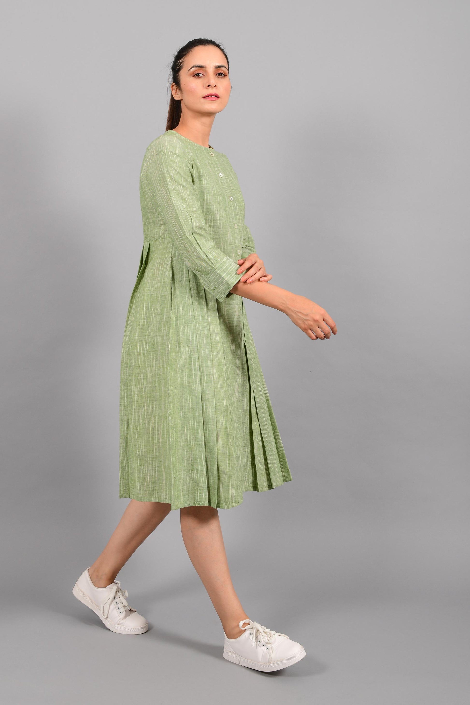 Stylised side pose of an Indian female womenswear fashion model in a space dyed olive green handspun and handwoven khadi cotton pleated dress-kurta by Cotton Rack.