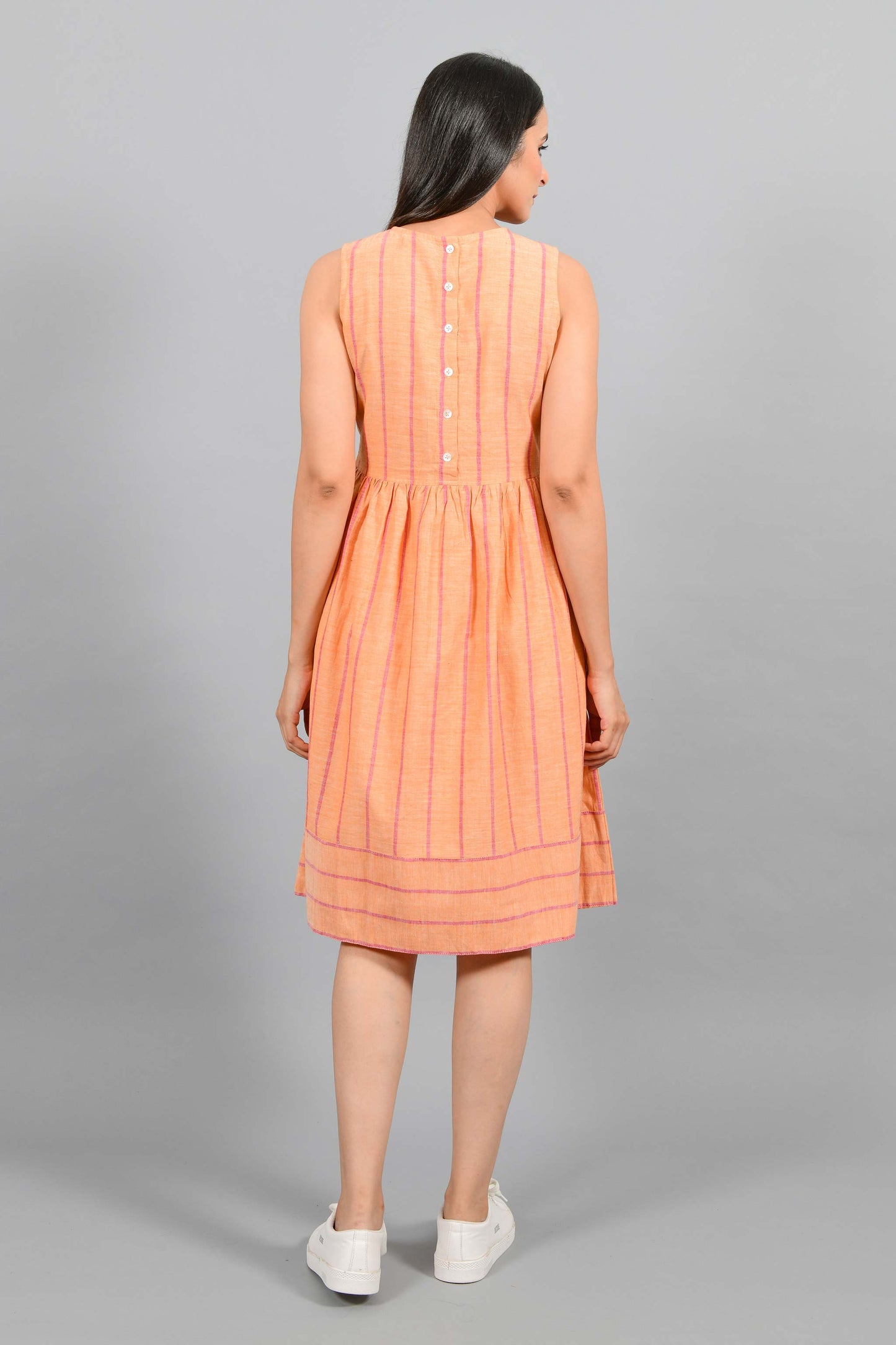 Back pose of an Indian female womenswear fashion model in a orange chambray handspun and handwoven khadi cotton with red stripes by Cotton Rack. The dress has gathers at waist. and the stripes are arranged in attractive manner.