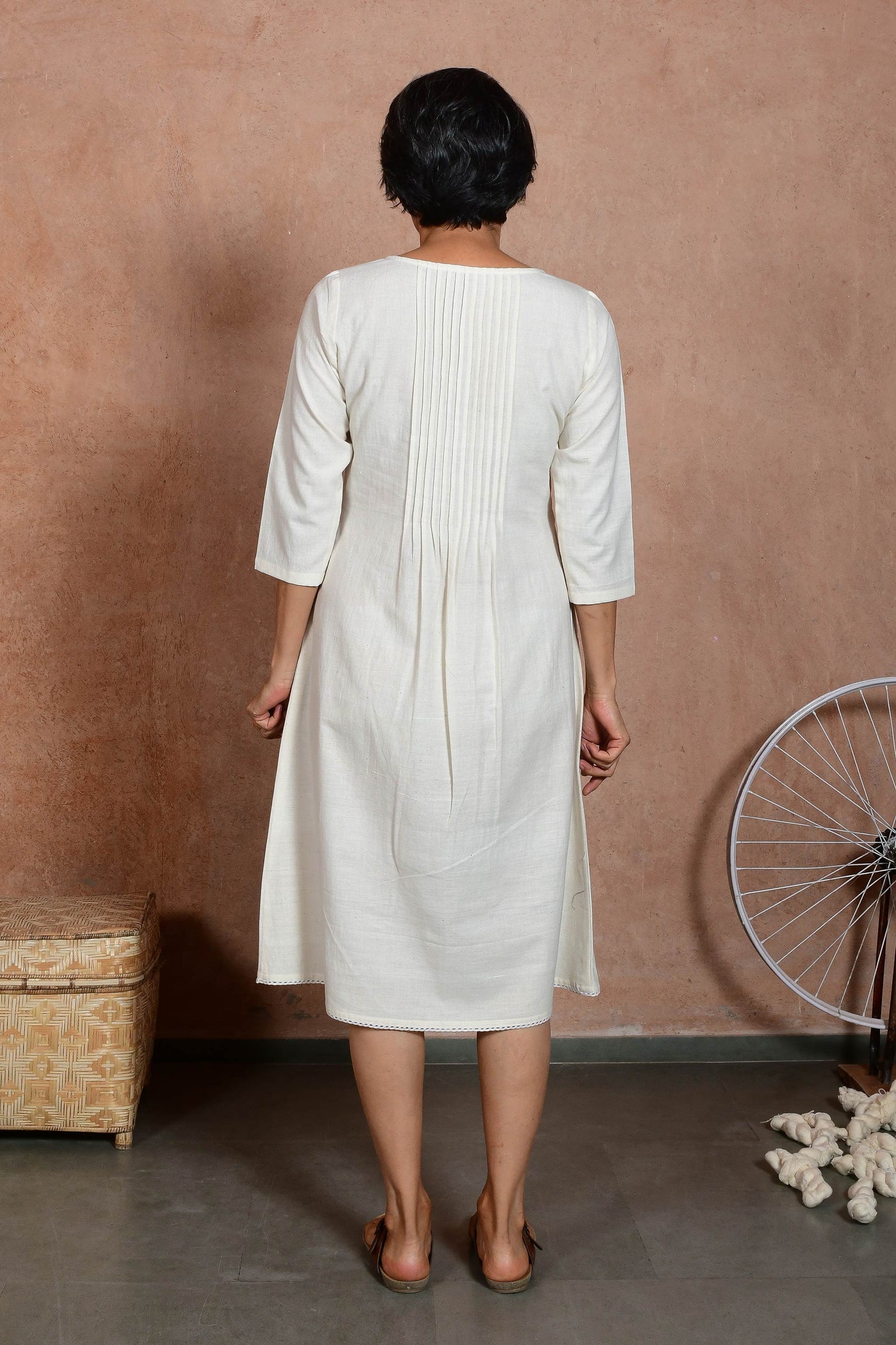 Back pose of a middle aged indian woman wearing an off white knee length dress with sleeves and knife pleat detail.