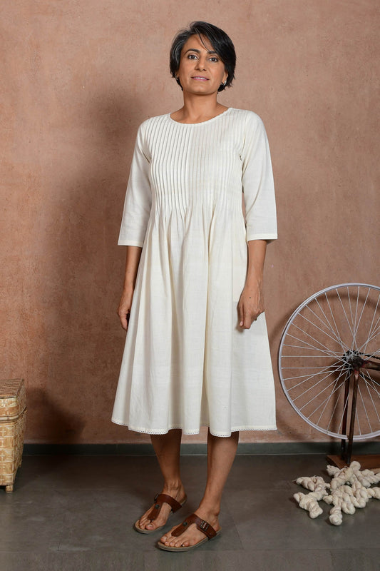 Front pose of a middle aged indian woman wearing an off white knee length dress with sleeves and knife pleat detail.