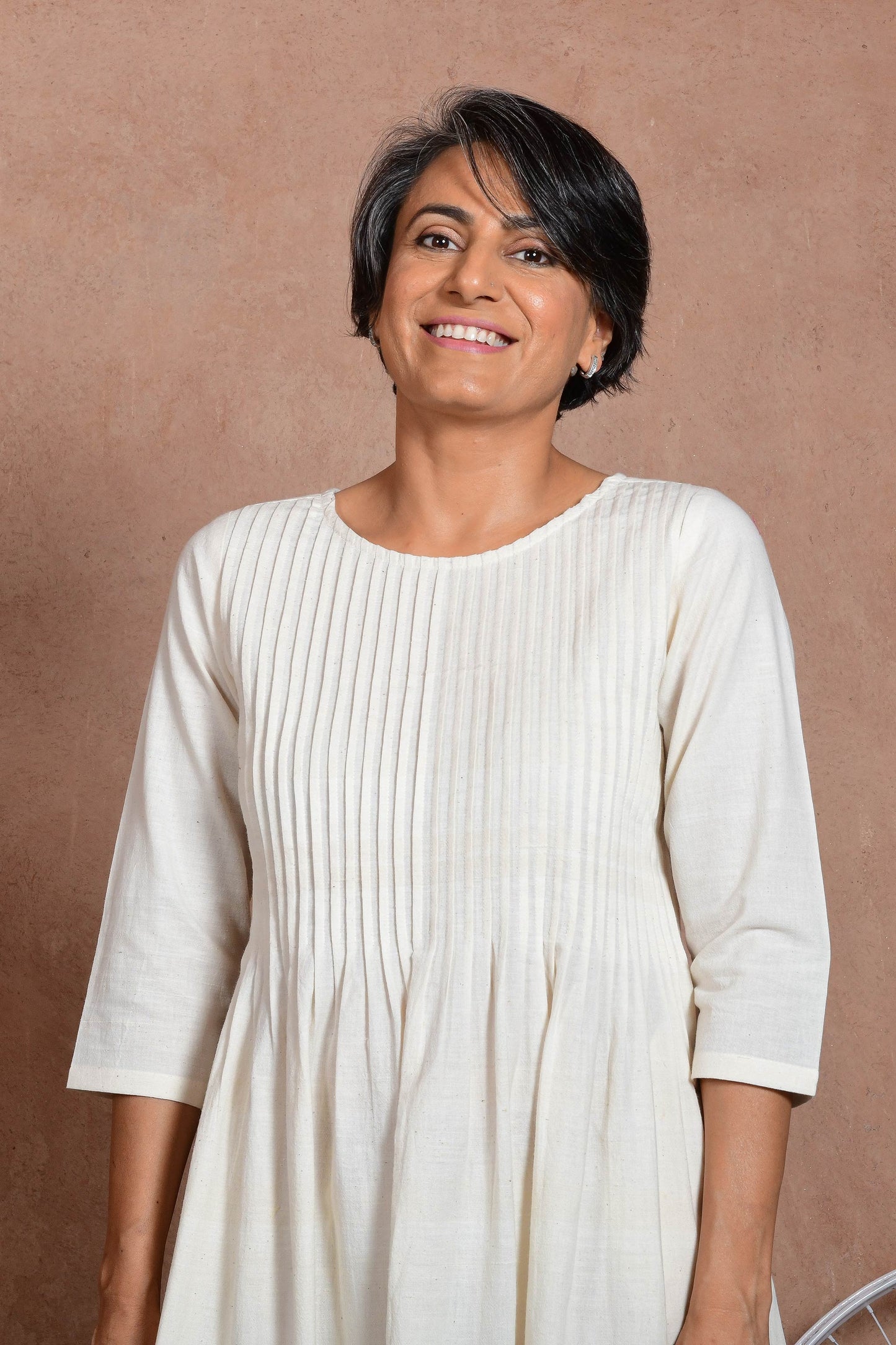 Close up of an upper half of middle aged indian woman wearing an off white knee length dress with sleeves and knife pleat detail.