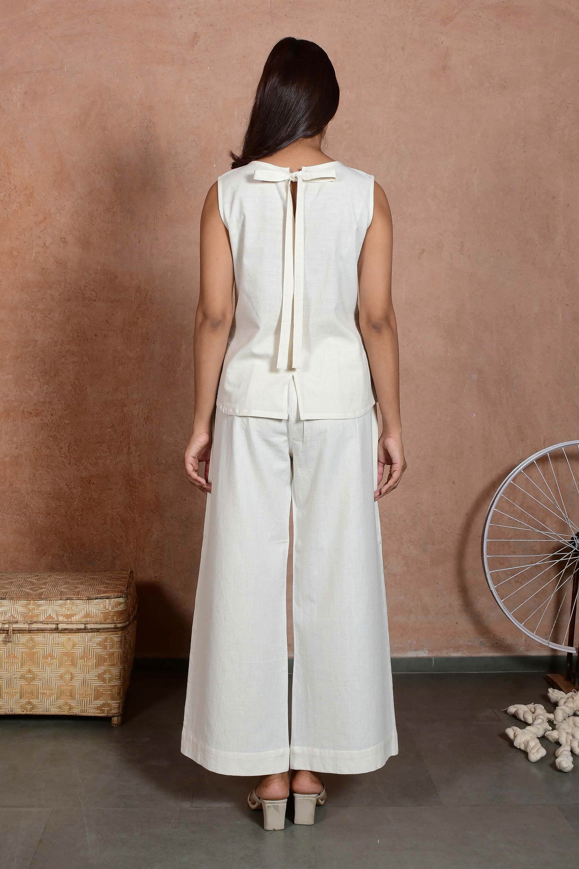 Back pose of young Indian model posing wearing off white sleeveless top with a bow tied at the neck and straight pants.