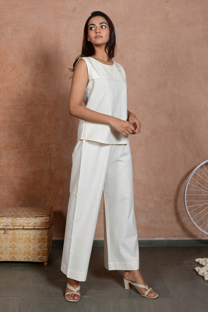A three quarters pose of young Indian model posing wearing off white sleeveless pleated top and straight pants.