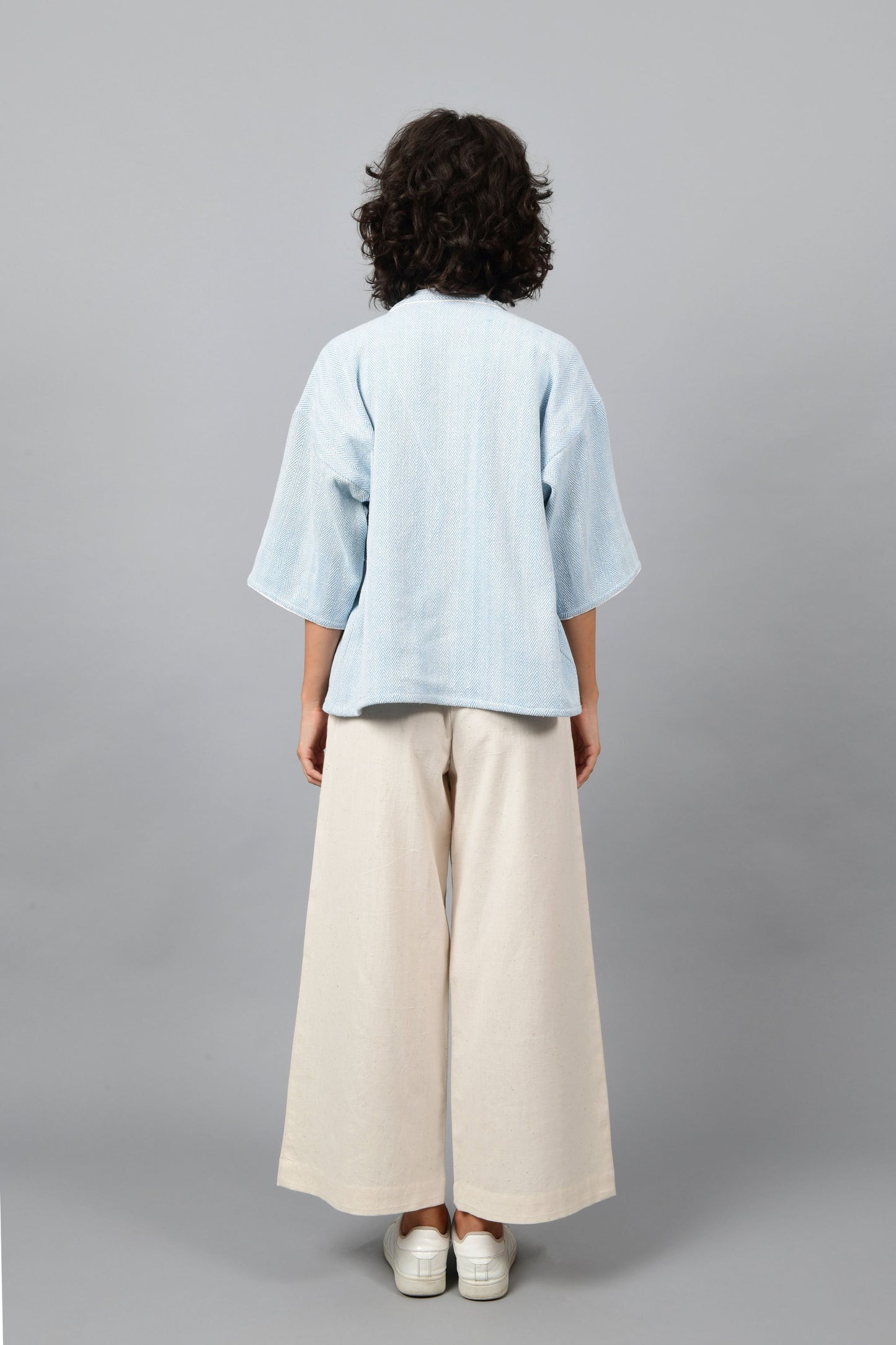Back of a model wearing anti-fit short blue shrug made in pointed twill with handspun and handwoven khadi cotton. Paired with off-white spaghetti, palazzos and white shoes.