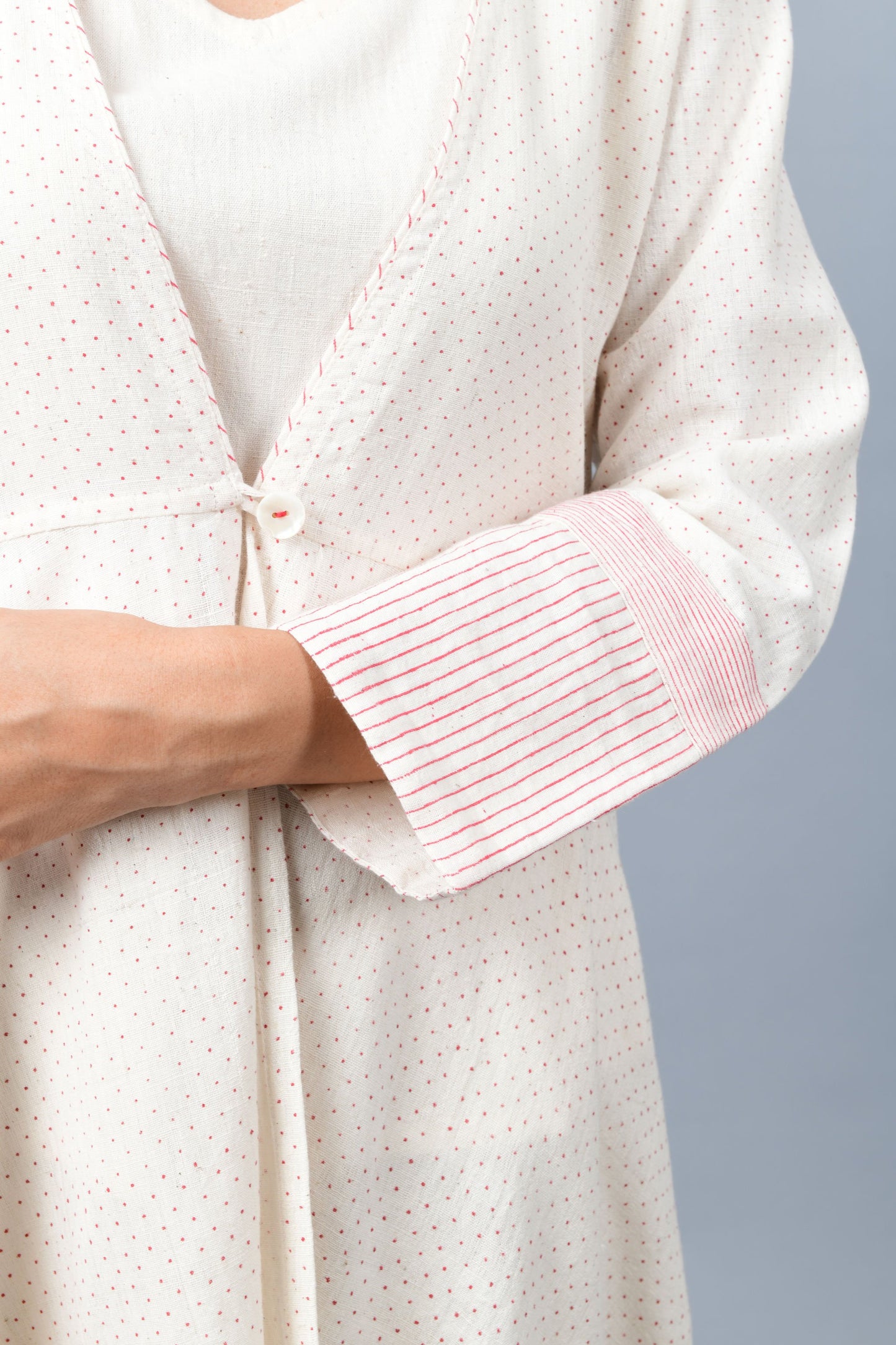 Close of a long flared off-white, natural khadi cotton shrug buttoned up in the front, hand block printed with fine red dots and red stripes on the sleeve hem.
