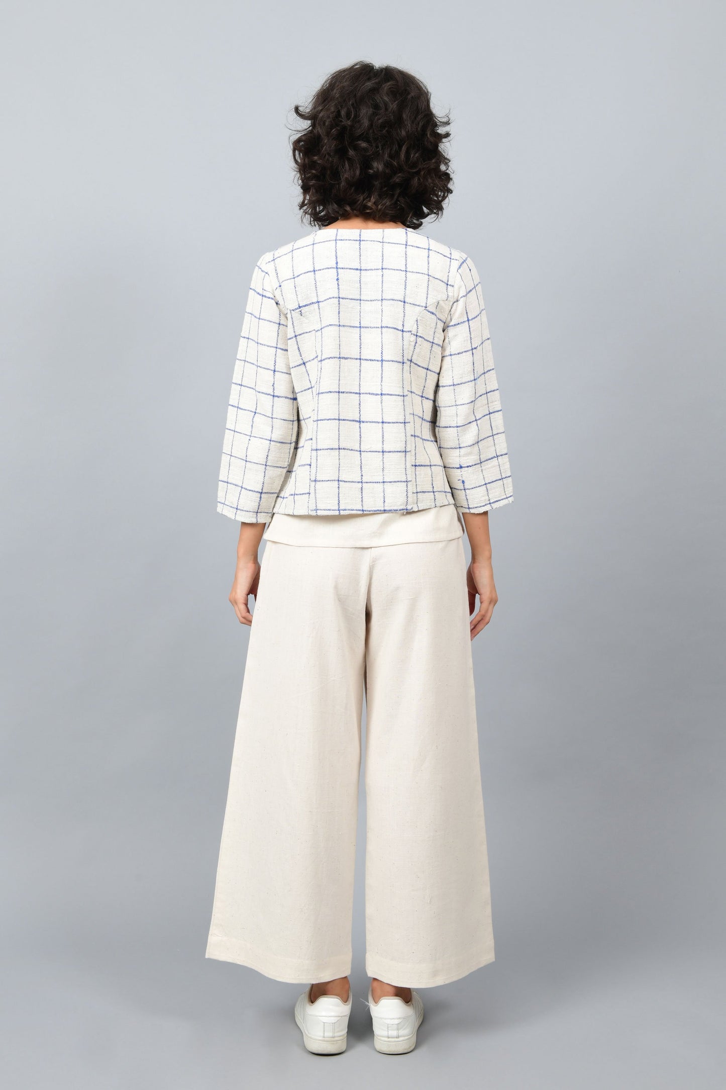 Model showing the back of open short jacket in thicker white handspun and handwoven khadi cotton with big blue checks over off-white spaghetti top and off-white palazzos paired with white sneakers.