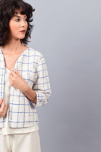 Close-up of model posing for the camera wearing open short jacket in thicker white handspun and handwoven khadi cotton with big blue checks over off-white spaghetti top and off-white palazzos paired with white sneakers.