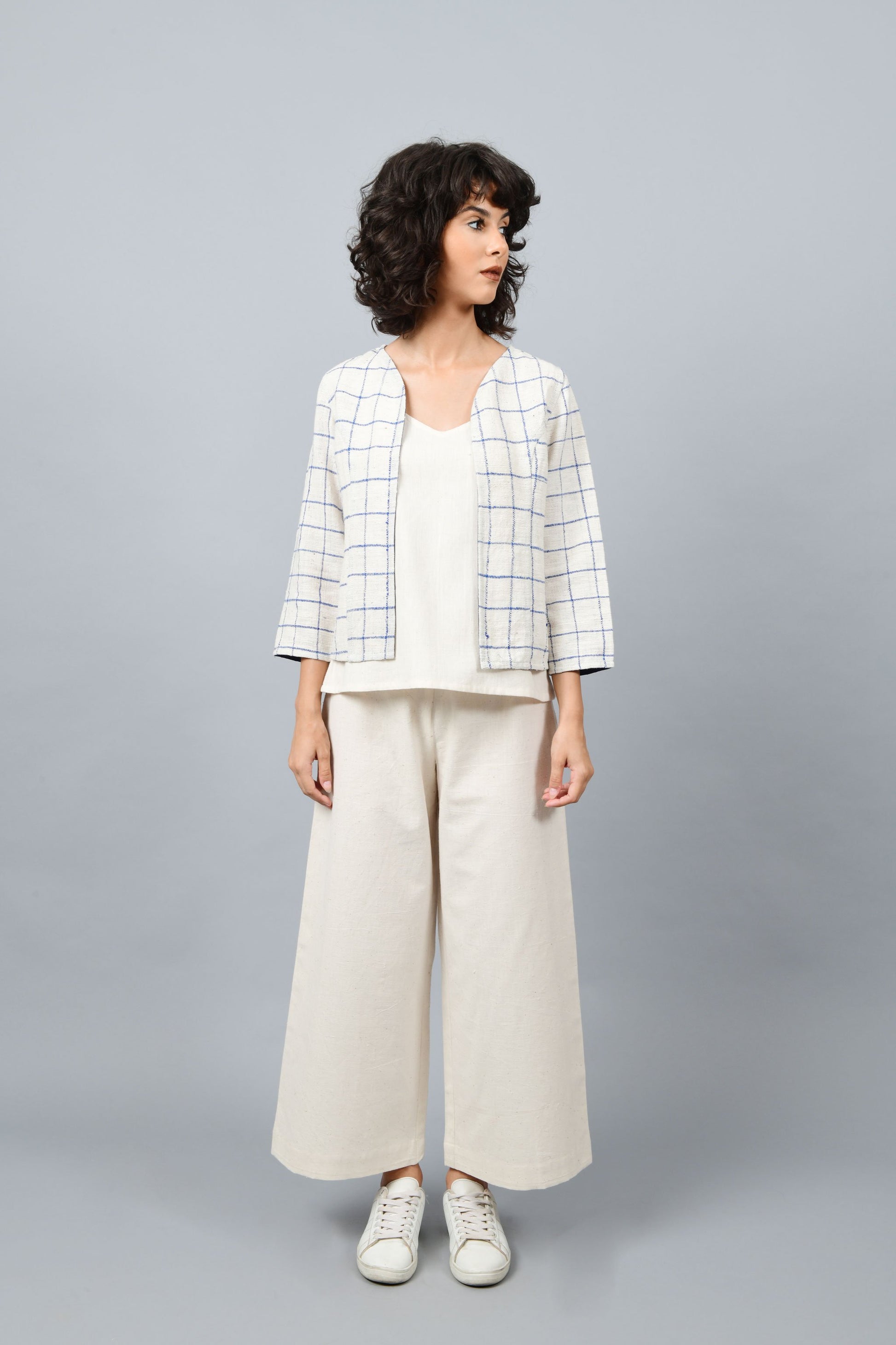 Model posing for the camera wearing open short jacket in thicker white handspun and handwoven khadi cotton with big blue checks over off-white spaghetti top and off-white palazzos paired with white sneakers.