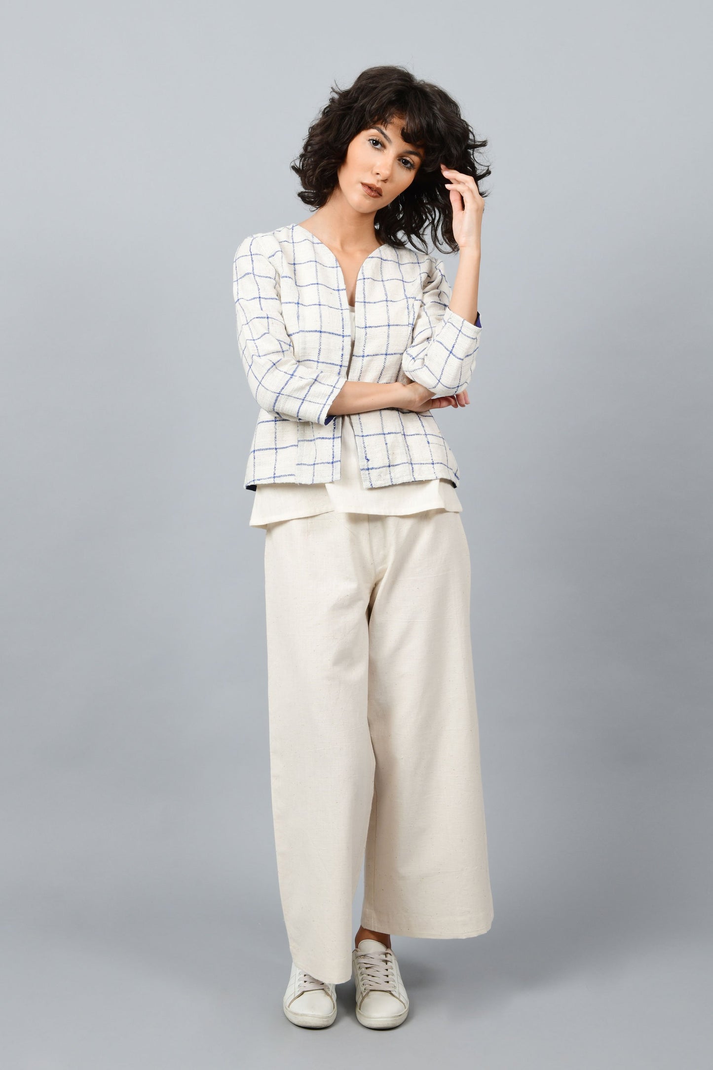 Brazilian Model posing for the camera wearing open short jacket in thicker white handspun and handwoven khadi cotton with big blue checks over off-white spaghetti top and off-white palazzos paired with white sneakers all made in India